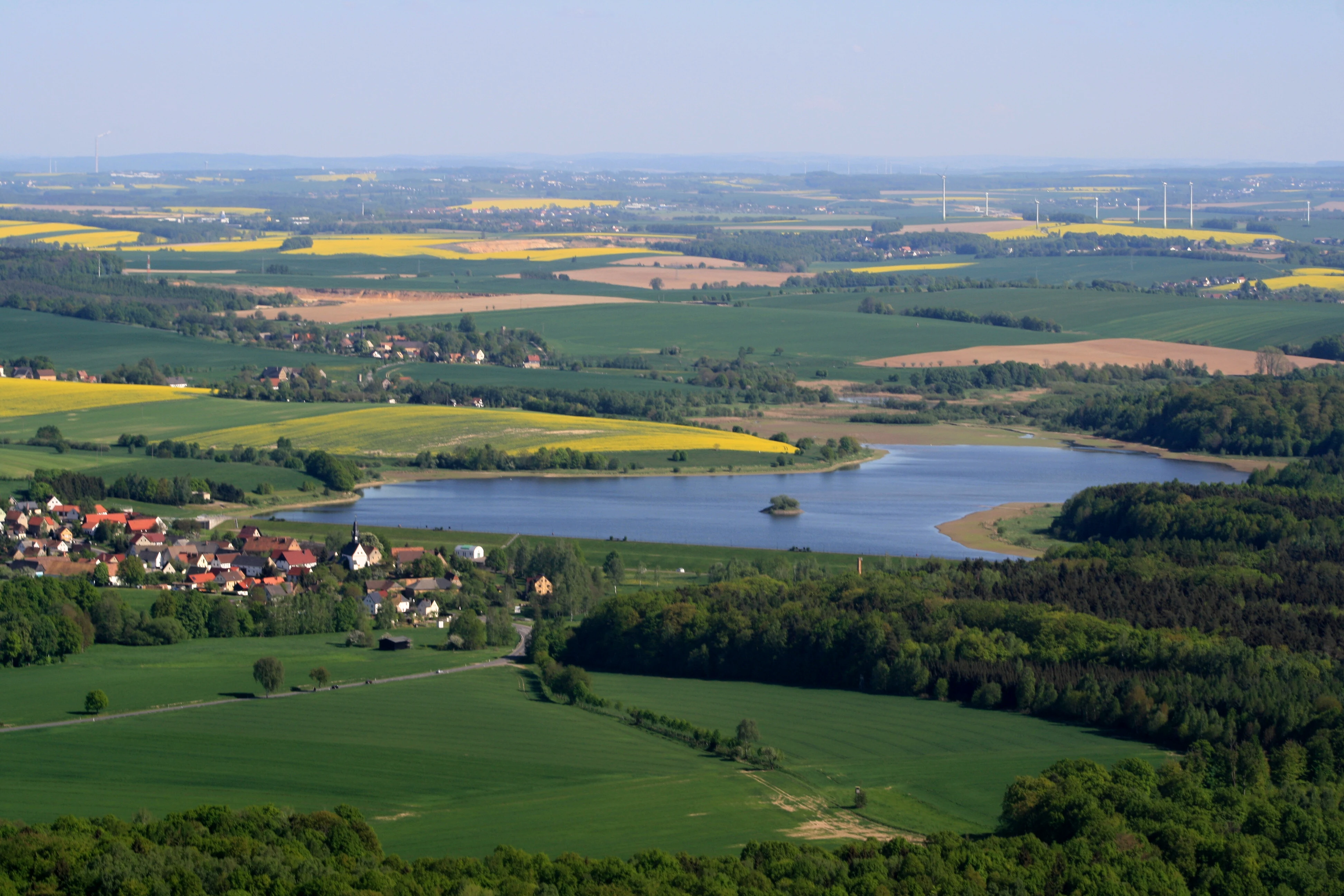 An elevated wide-angle view of a reservoir surrounded by agricultural fields, patches of forest, and a small town.