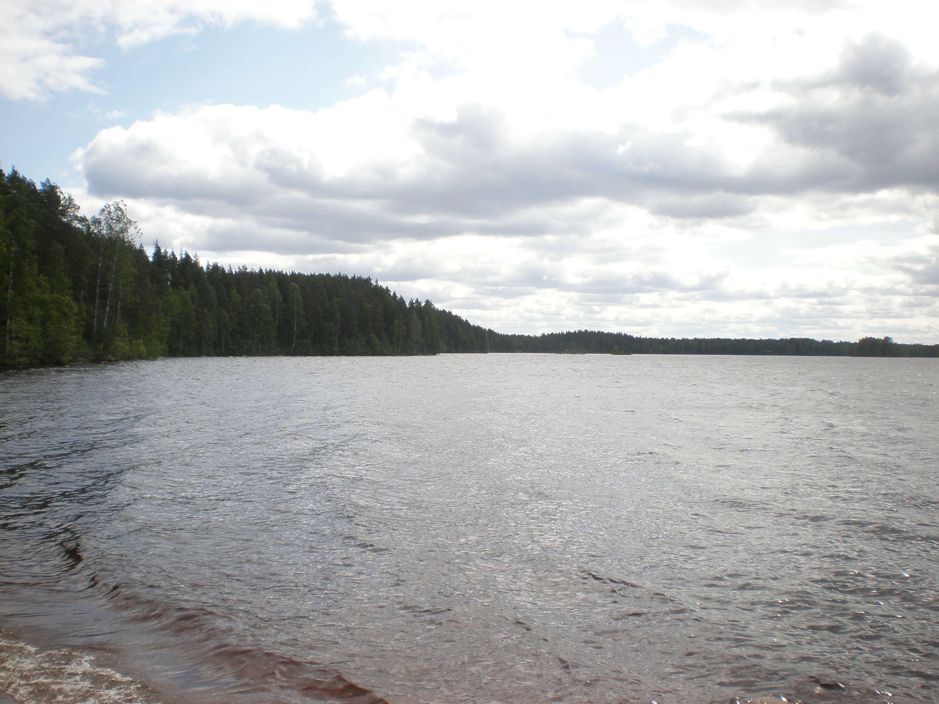 Calm lake with pebbly shore, forested shoreline, and partly cloudy sky