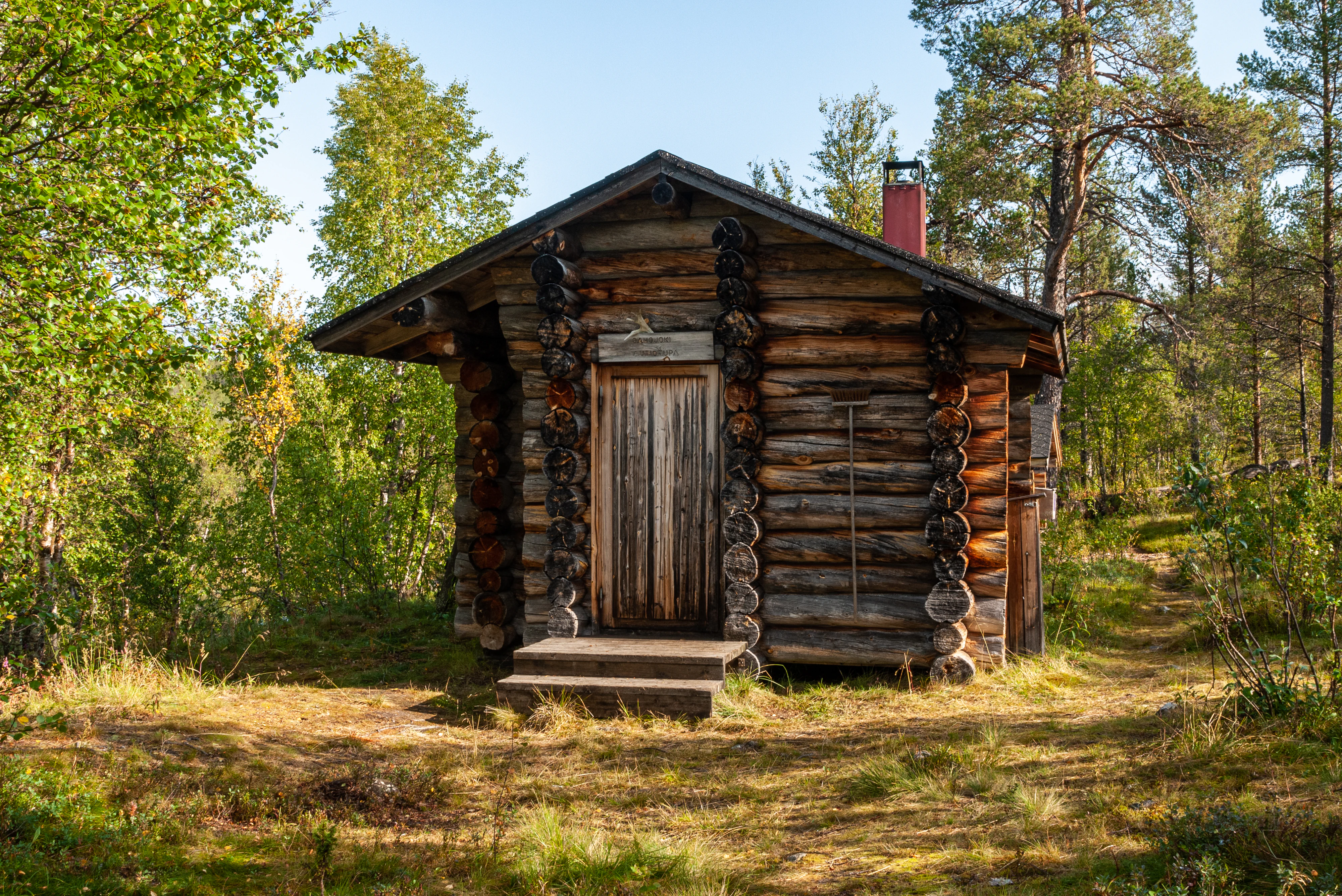 Wooden log cabin with stone steps and stone pillars, surrounded by trees and grassy terrain
