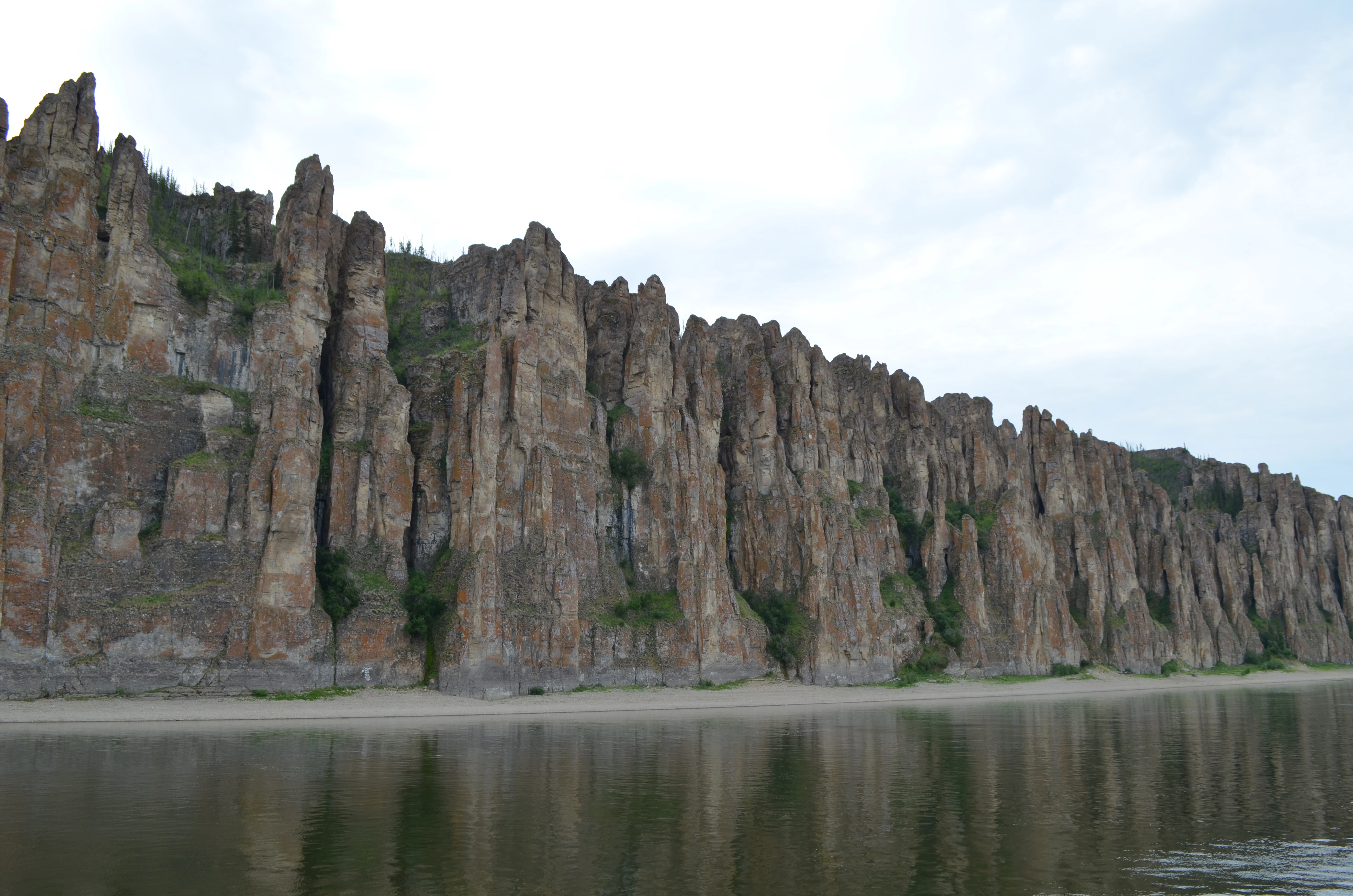 A long, sweeping view of the vertical rock pillars at Lena Pillars National Park, with a sandy riverbank at their base and the reflection of the rock face in the water of the Lena River.