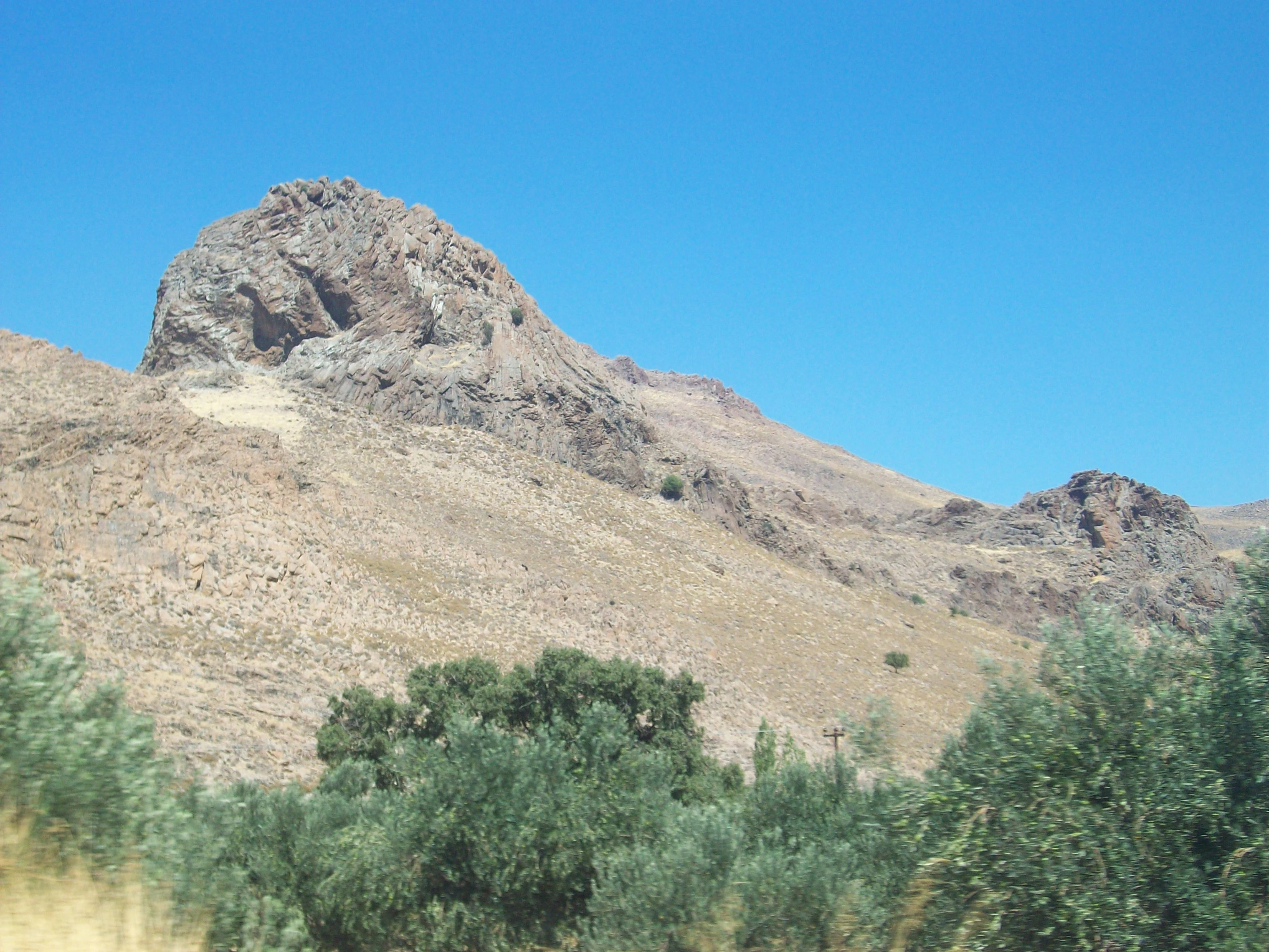 A rocky, arid volcanic lava dome rises above a sparse landscape of green trees under a clear blue sky on the island of Lesvos.