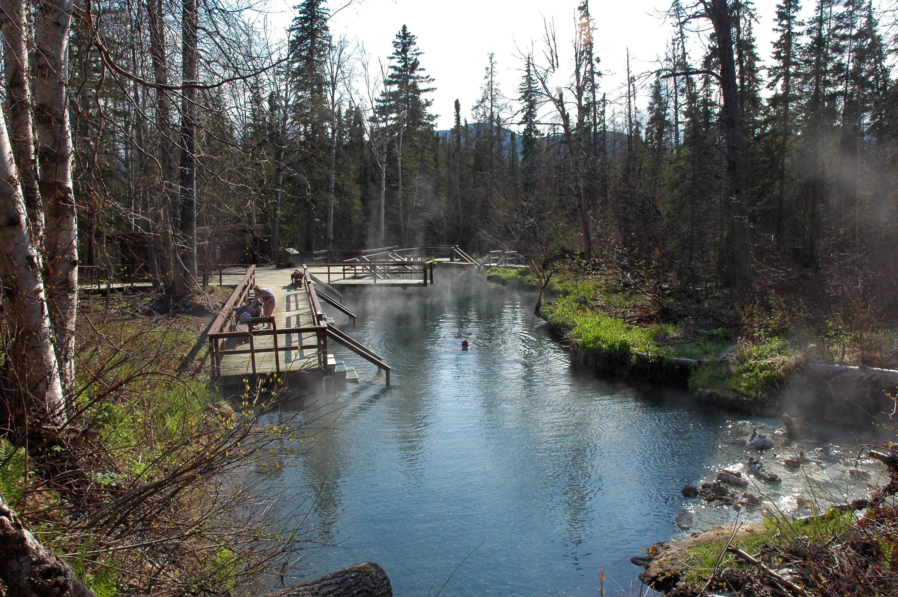 A natural thermal pool with steam rising from the surface, flanked by a wooden boardwalk and surrounded by dense boreal forest.