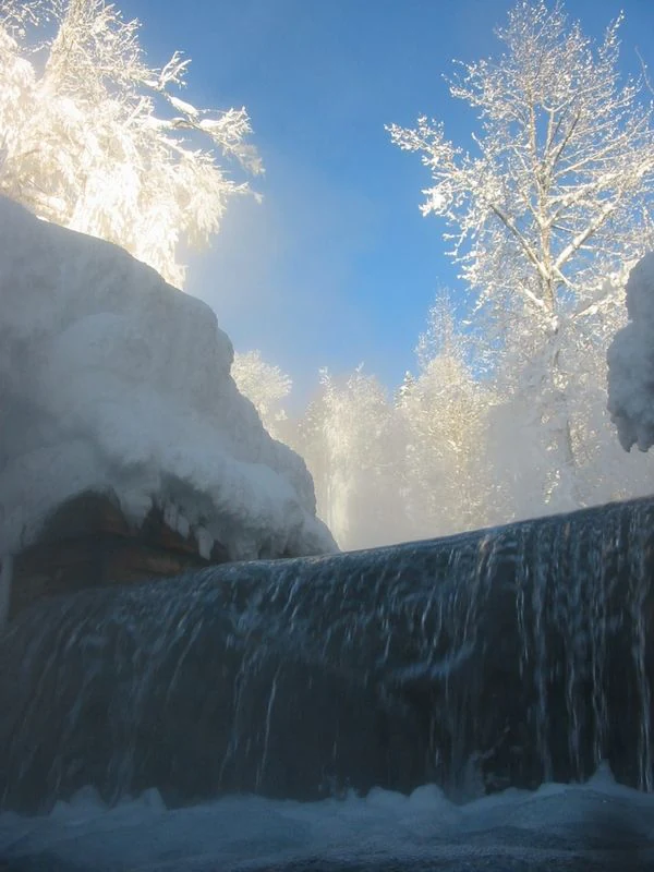 A low-angle view of a small thermal waterfall cascading over a ledge, surrounded by snow-covered trees and heavy frost in a winter forest.