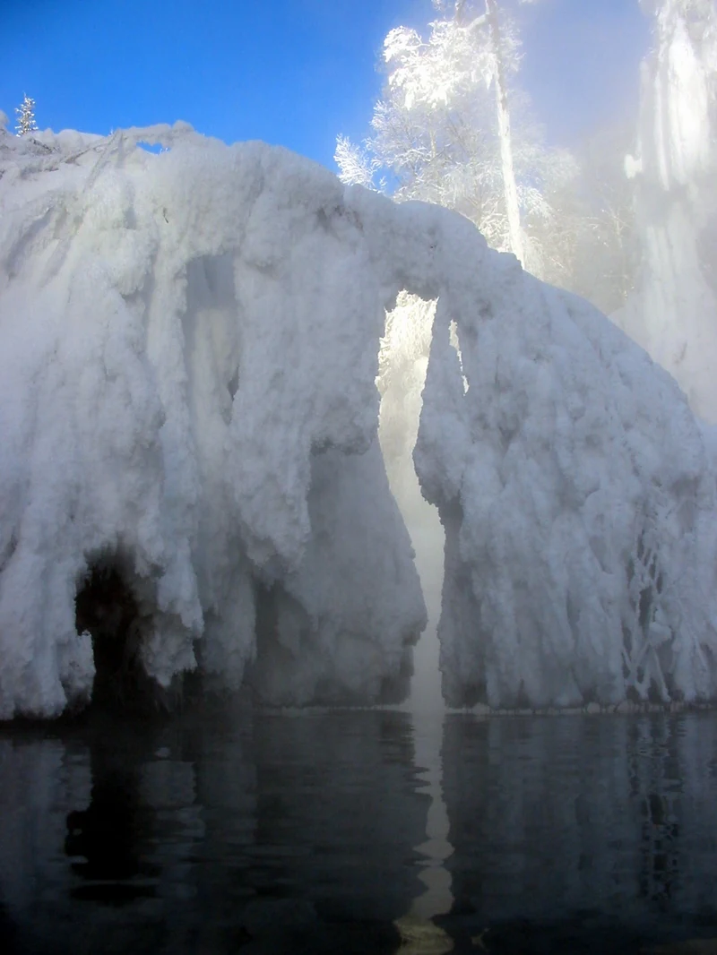 A natural hot spring pool in winter with heavy ice and snow formations hanging over the water, creating a steam-filled gap against a clear blue sky.