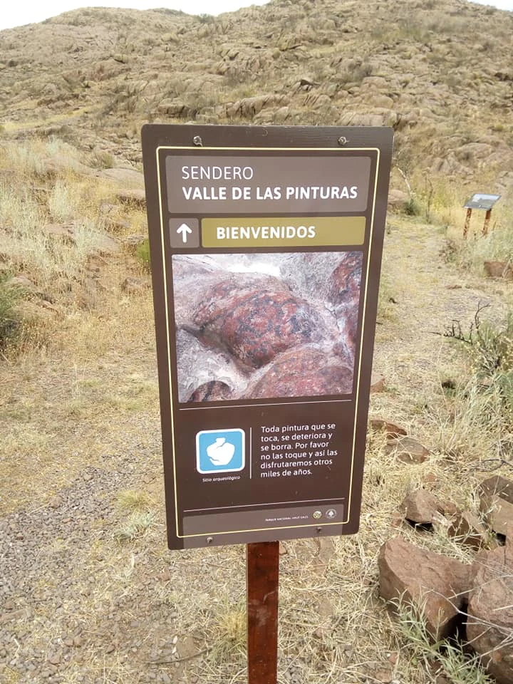 Brown trailhead sign reading 'Sendero Valle de las Pinturas' and 'Bienvenidos', showing an image of rock art. Arid landscape with rocky hills in background.