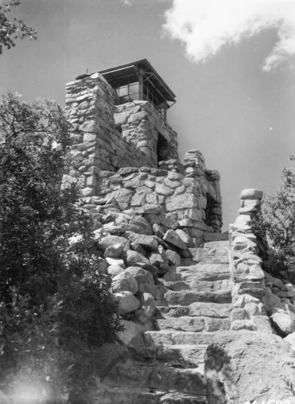 A vintage black and white photograph of a stone fire lookout tower with exterior stone stairs leading up to the structure.