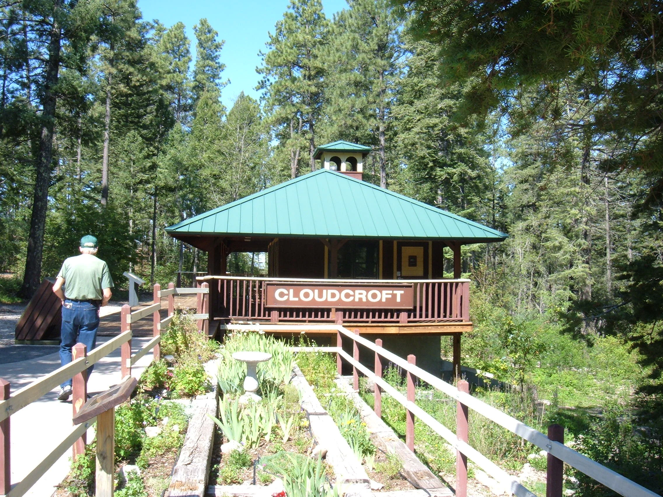 A small wooden building with a green metal roof and a sign reading Cloudcroft, surrounded by a pine forest and wooden railed walkways.