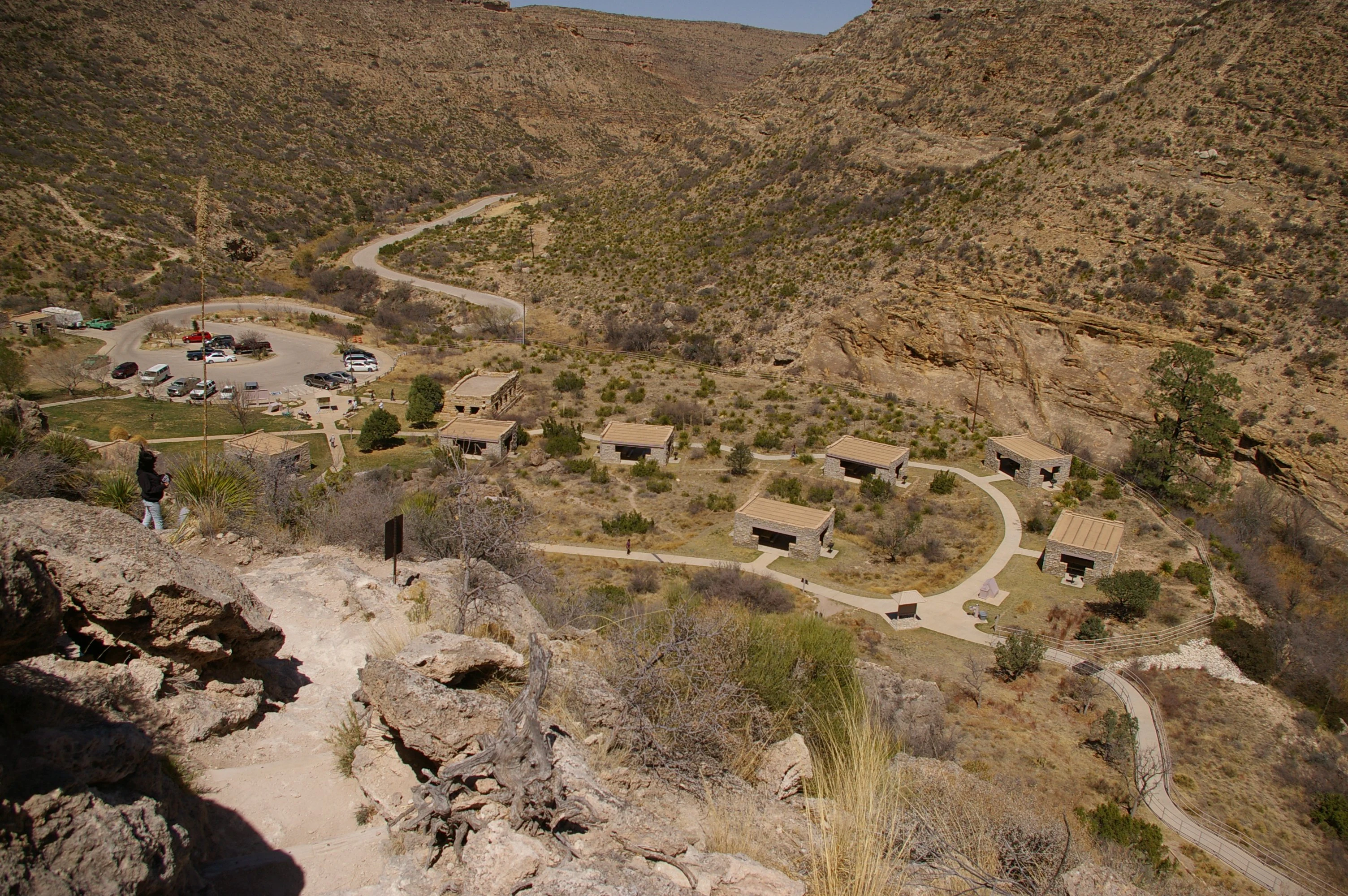 A high angle, wide shot looking down into a desert canyon at a day use area containing several stone picnic shelters, a parking lot with vehicles, and paved walkways, surrounded by arid hills.