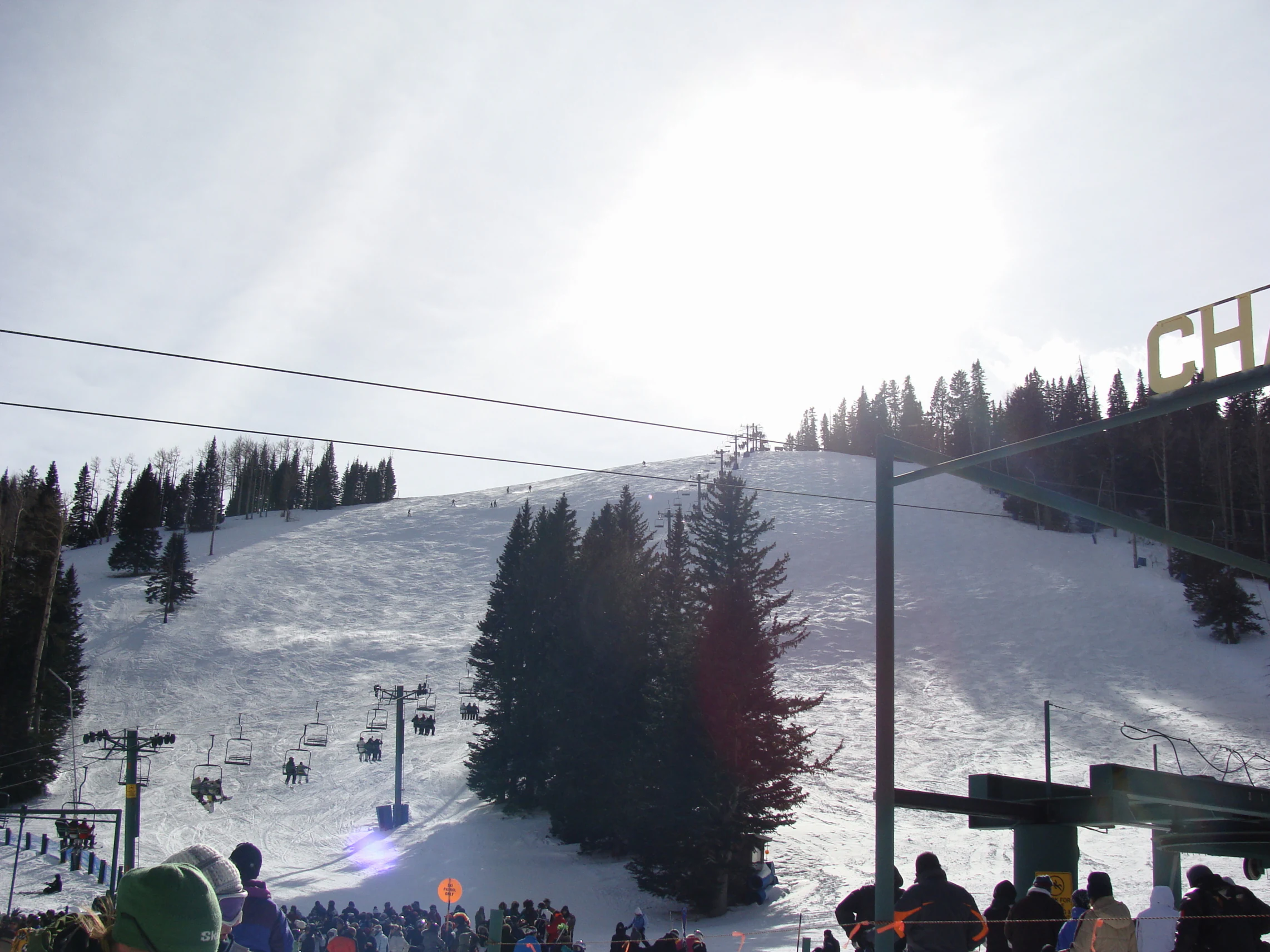 A wide shot looking up a snowy mountain slope at a ski resort with visible ski lifts, pine trees, and a group of people gathering at the base.