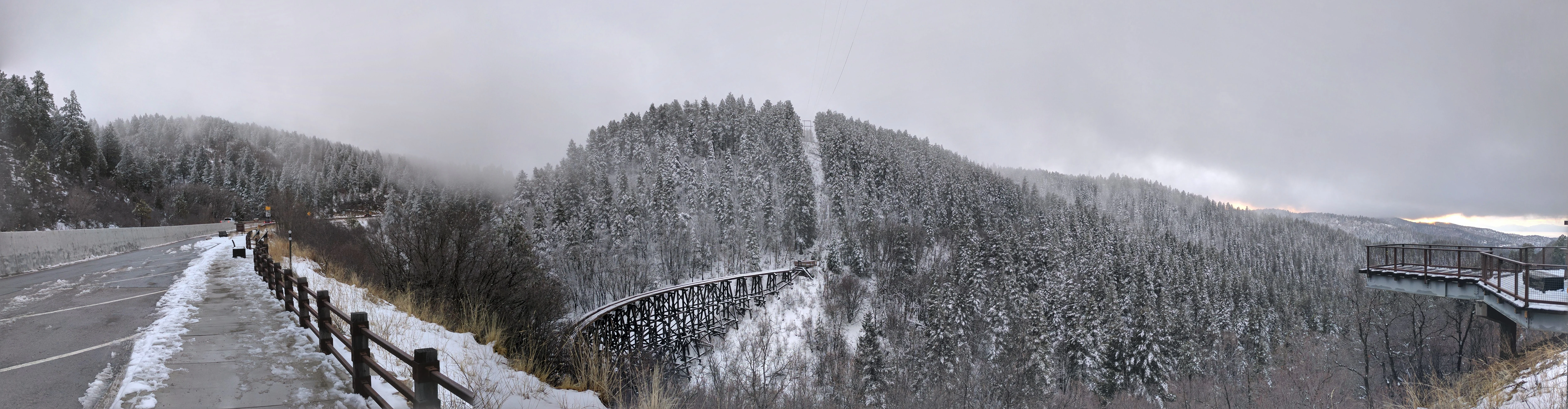 A wide panoramic view of a wooden trestle bridge spanning a deep, snow-covered forest canyon, viewed from a roadside observation area on a cloudy day.