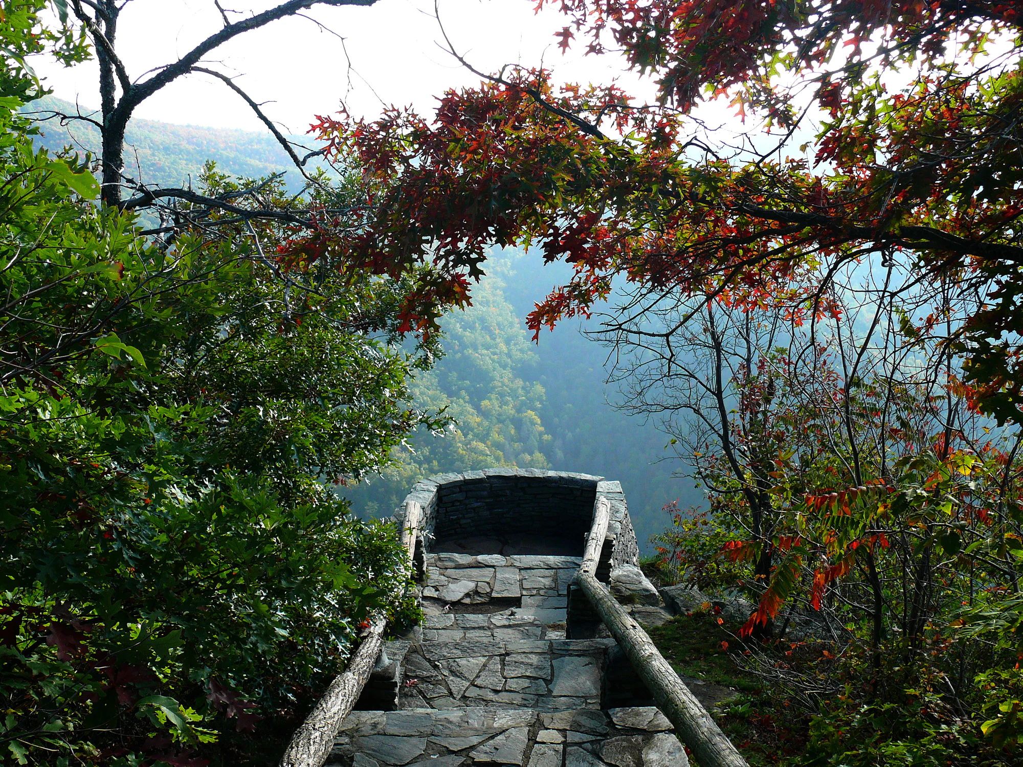 A stone-paved overlook with a simple wooden railing at the edge of a cliff, framed by overhanging trees with early autumn foliage, overlooking a deep, forested mountain gorge.