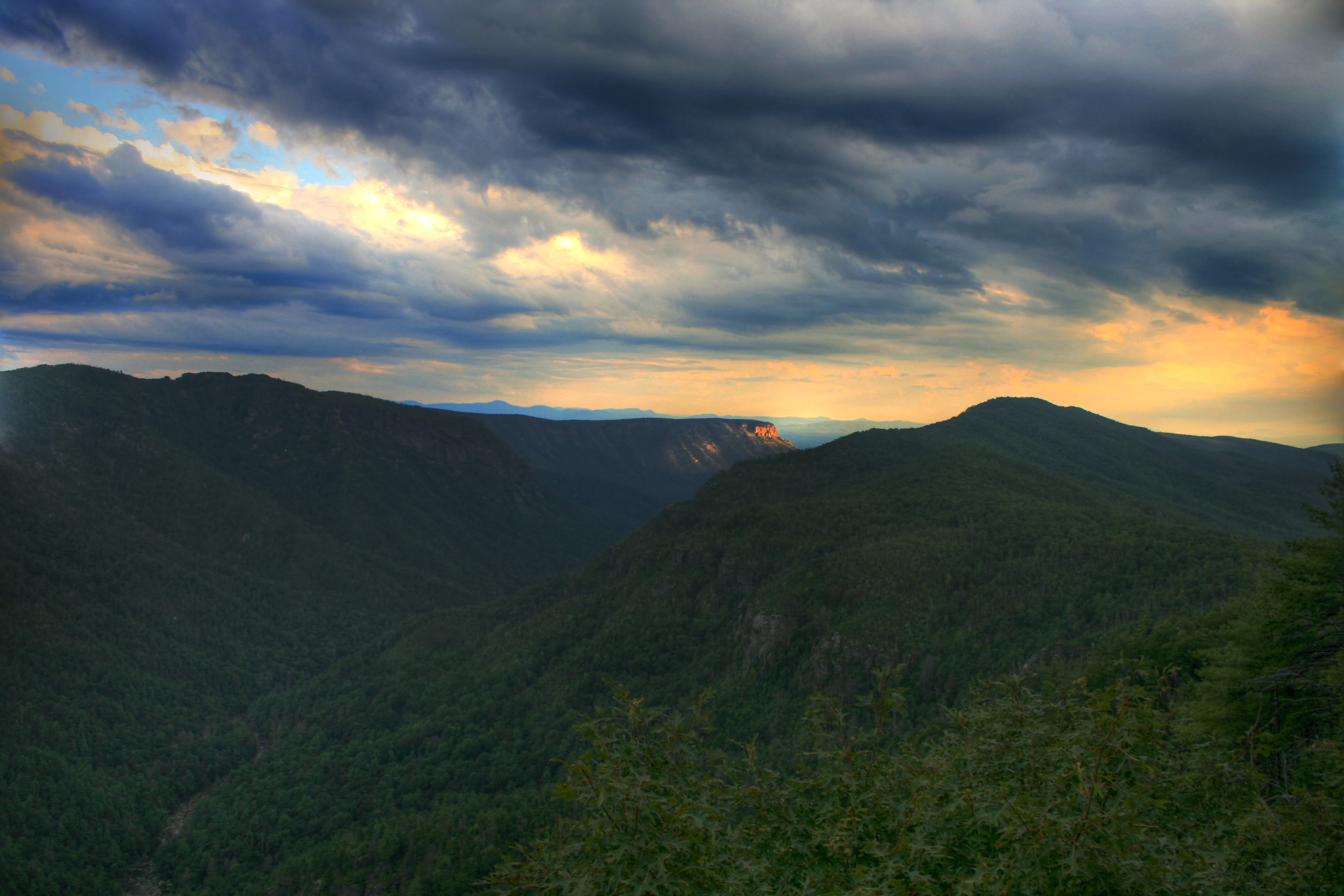 A wide, dramatic landscape view of forested mountains and a deep valley under a moody, cloud-filled sunset sky.