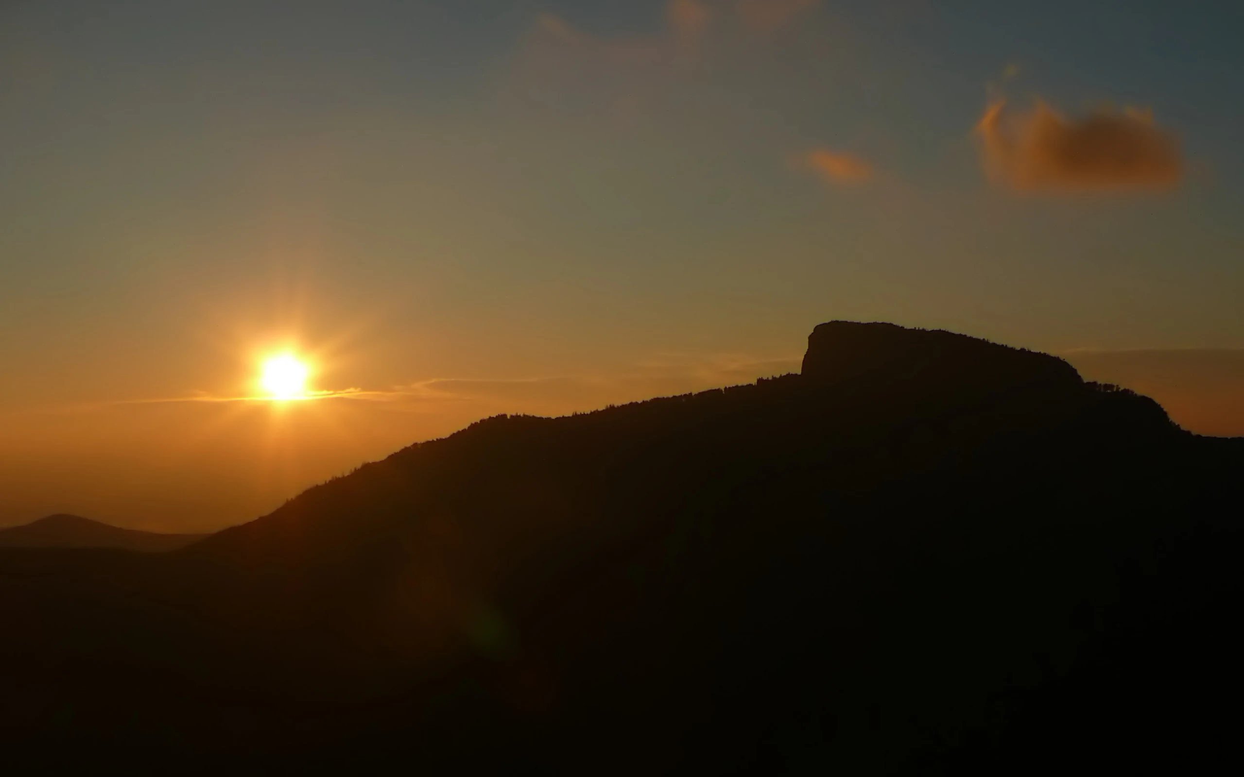 A backlit silhouette of a rocky mountain ridge at sunrise, with the sun low in the sky, creating a warm glow against the dark terrain.