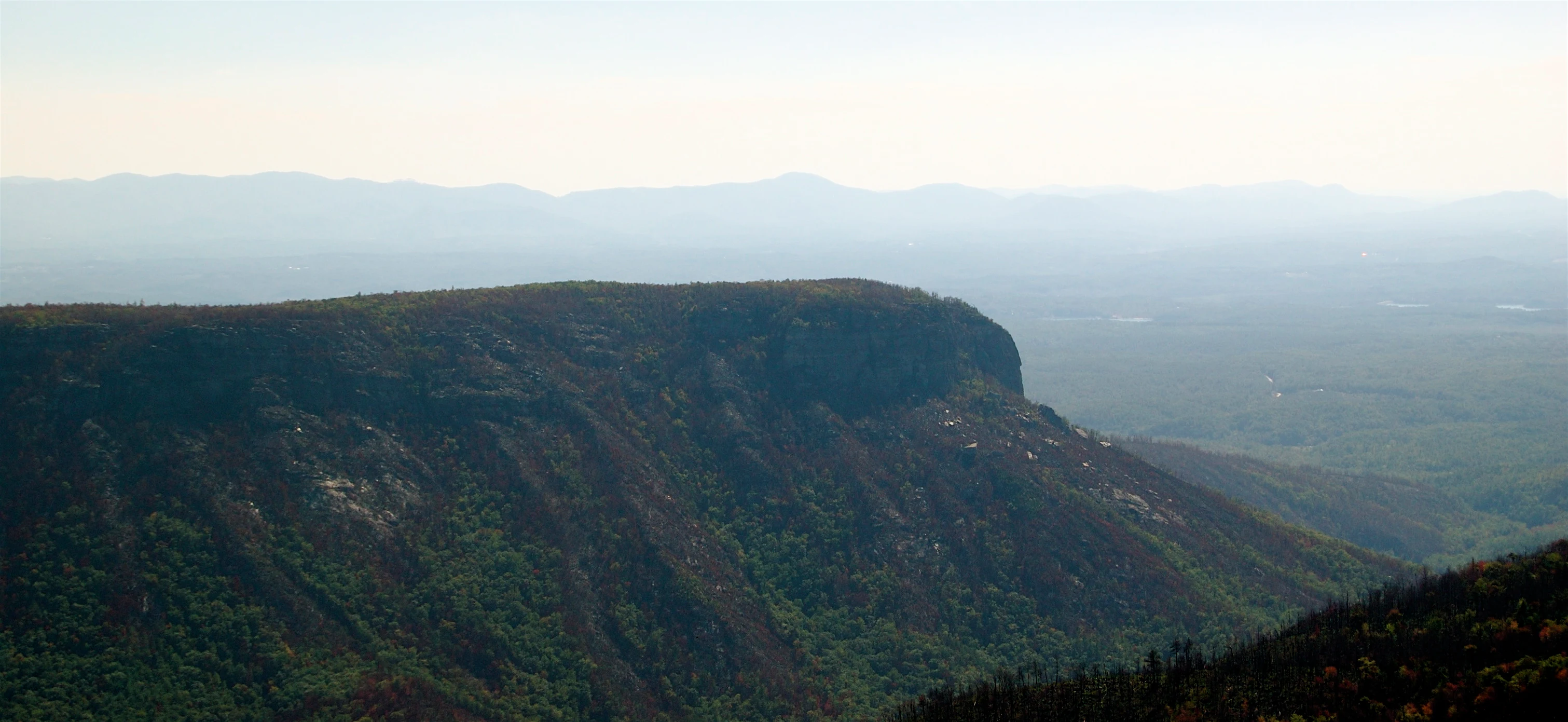 A high-angle scenic view of a prominent rocky ridge and deep gorge, with dense forested slopes and hazy distant mountains in the background.