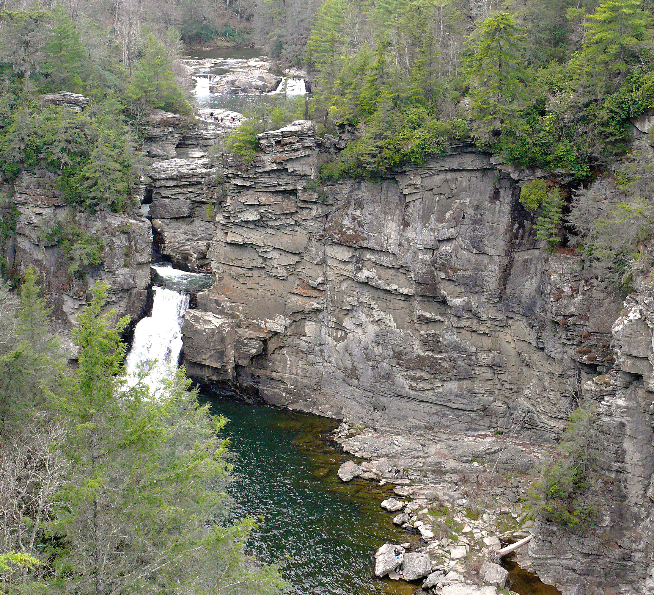 A multi-tiered waterfall flows down through a deep, rocky canyon surrounded by dense pine and hardwood forest.