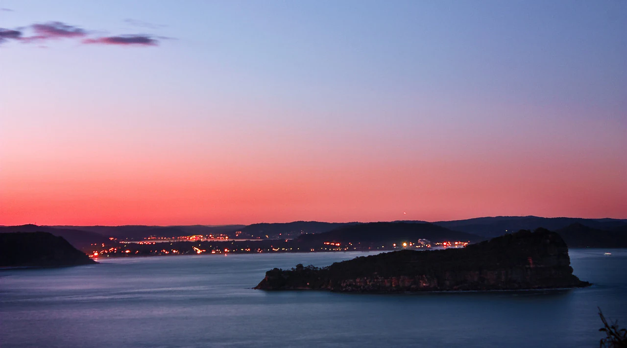 A wide view of Lion Island silhouetted against a vibrant pink and purple sunset sky over the water, with distant city lights visible on the horizon.