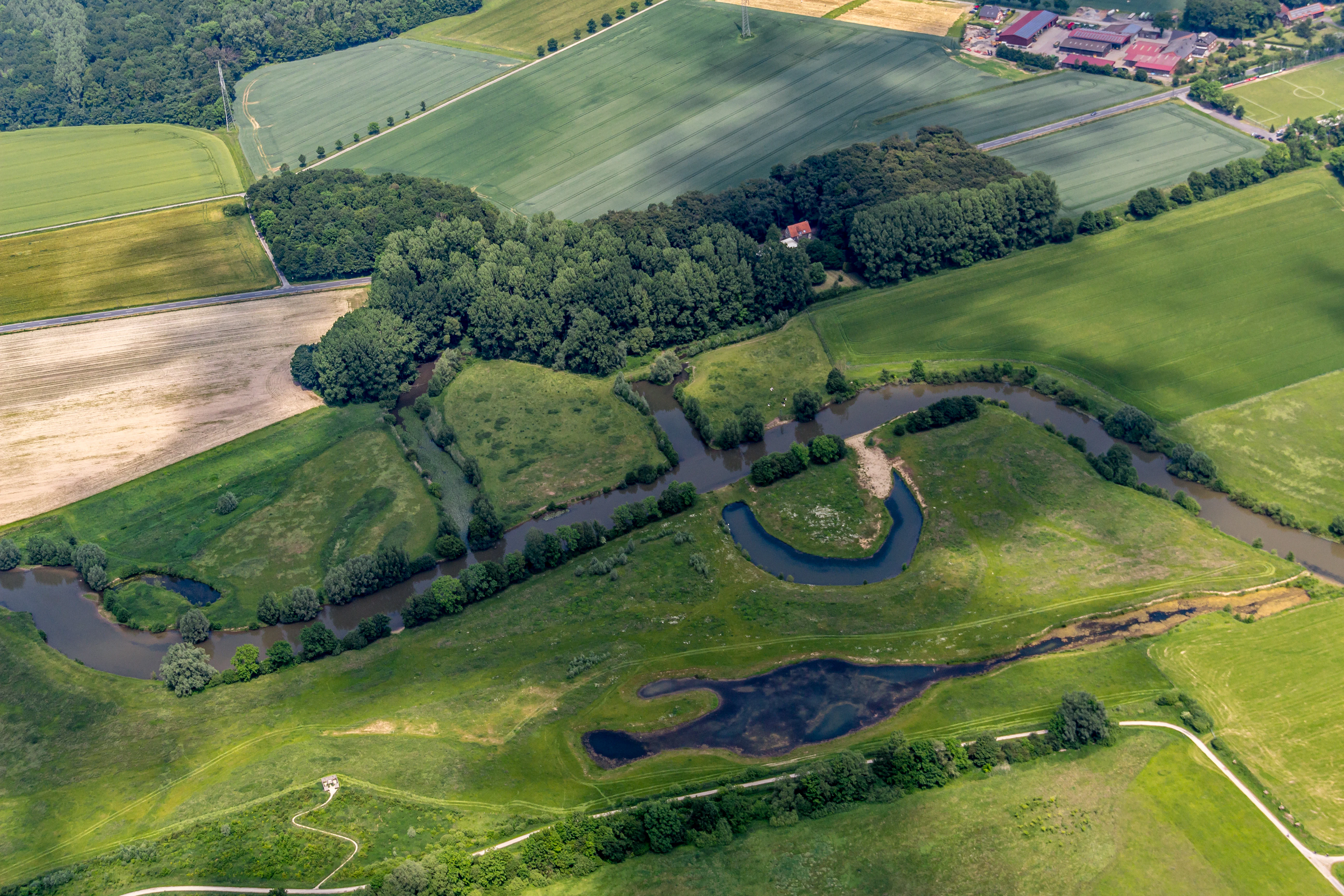 Aerial perspective of a meandering river section flowing through green meadows and patches of forest, with agricultural fields in the background.