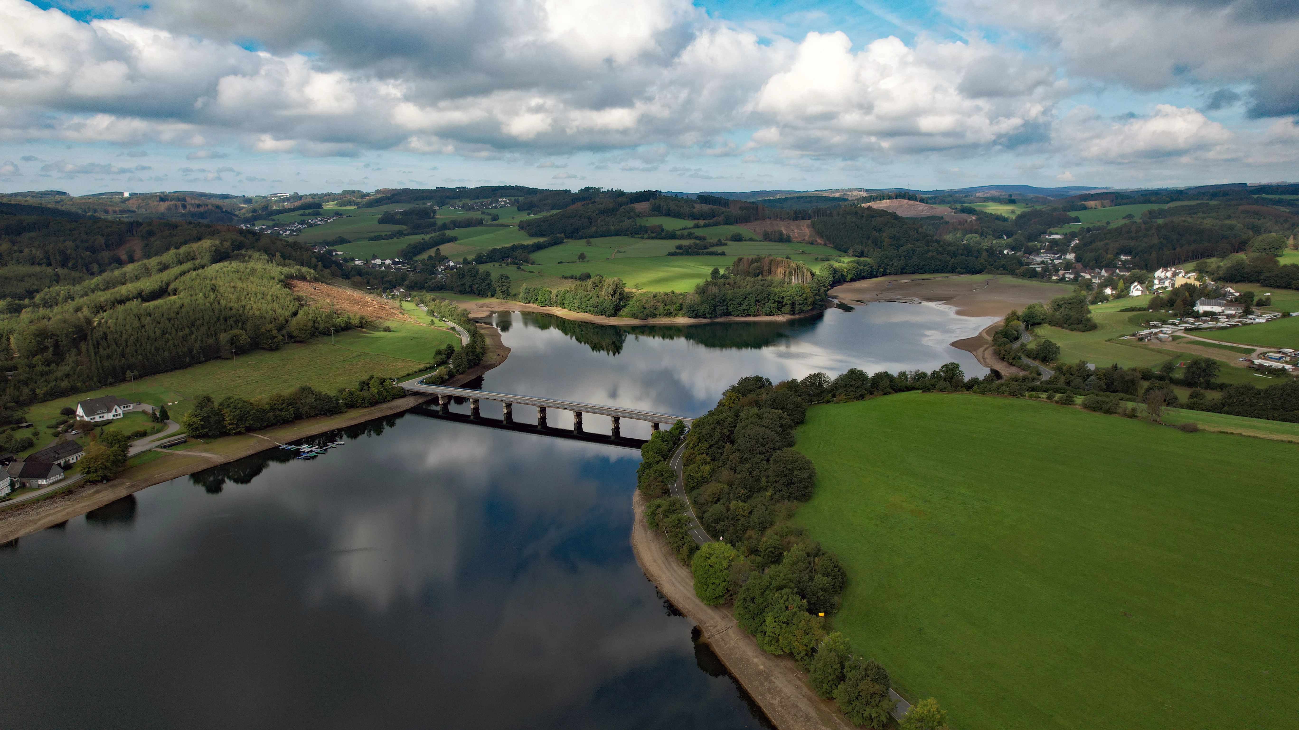 An aerial shot of a long reservoir bridge spanning a calm body of water, surrounded by green fields, pastures, and rolling hills under a cloudy sky.