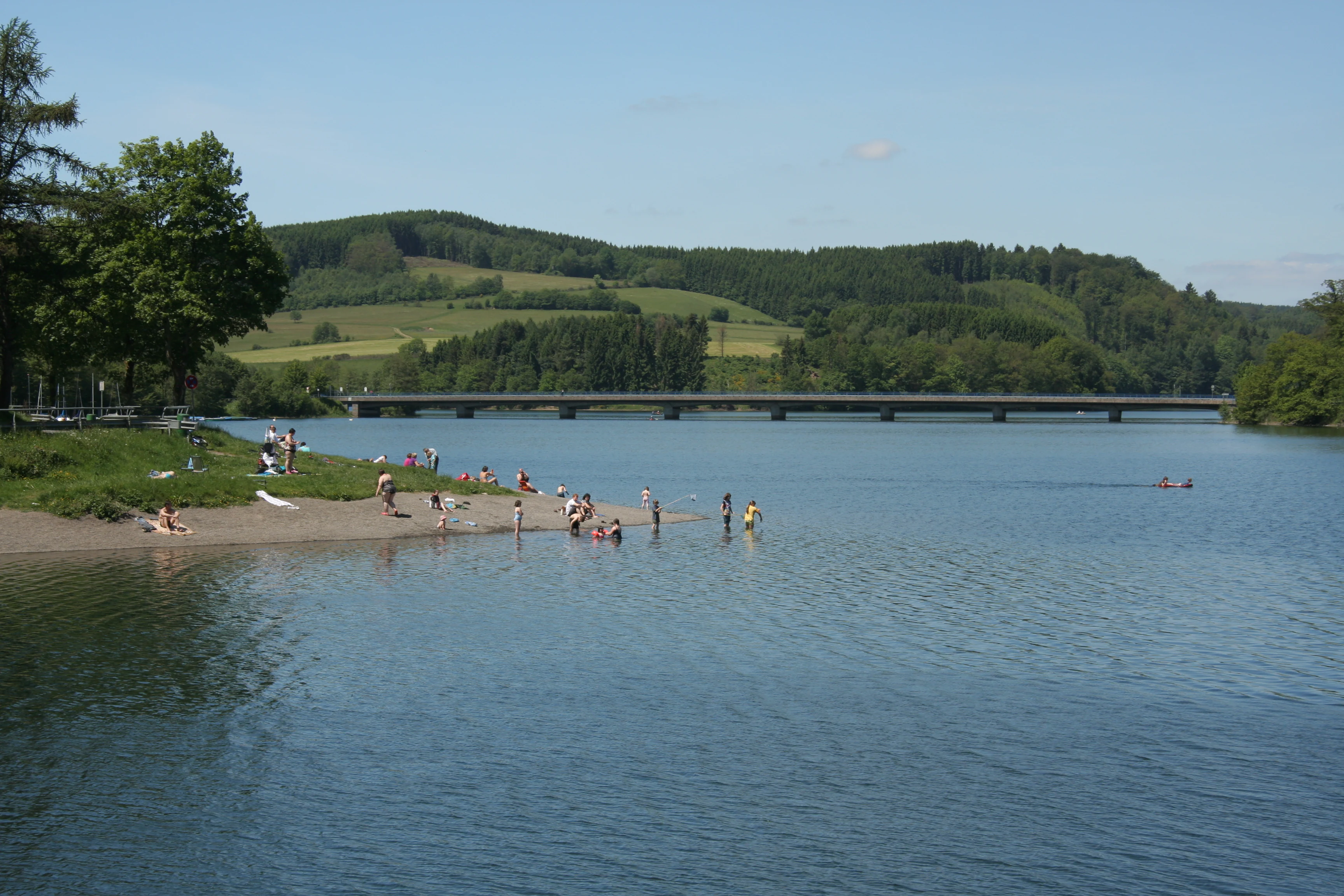 A group of people standing in and near the water on a small sandy beach at the edge of the Listertalsperre reservoir, with a long bridge visible in the background against rolling green hills.