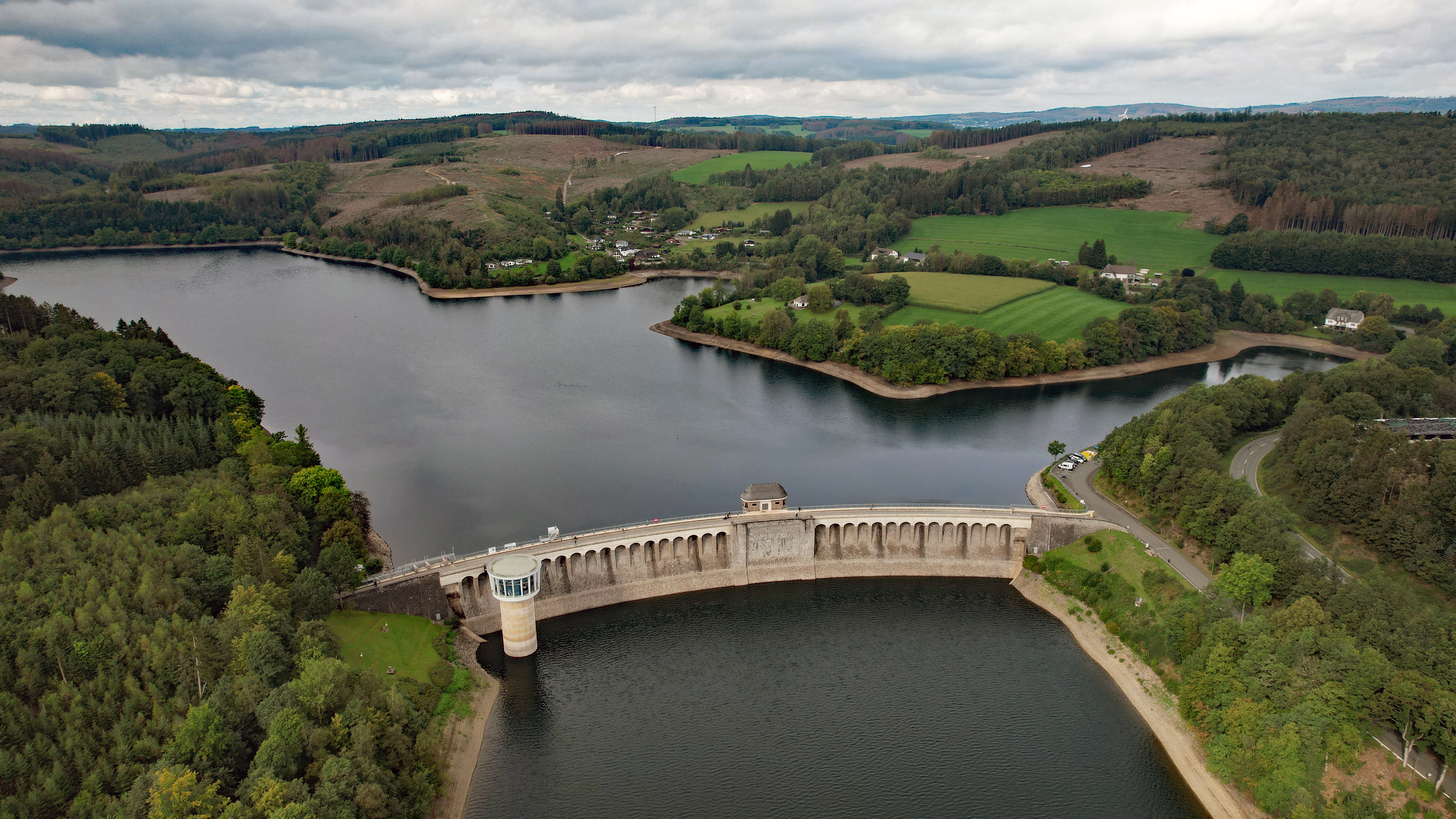 An aerial shot of a large curved concrete dam spanning across a wide reservoir, surrounded by lush green hills, forests, and fields under a cloudy sky.