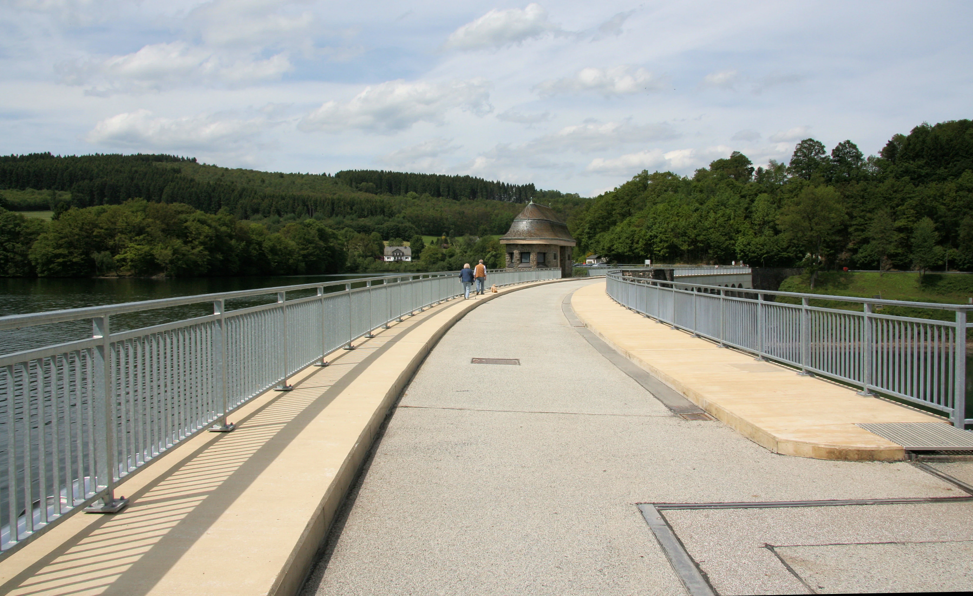A paved path runs across the dam wall of a reservoir, flanked by metal railings on both sides. In the distance, a small dome-roofed structure sits on the dam, with people walking towards it. Green forested hills surround the water.