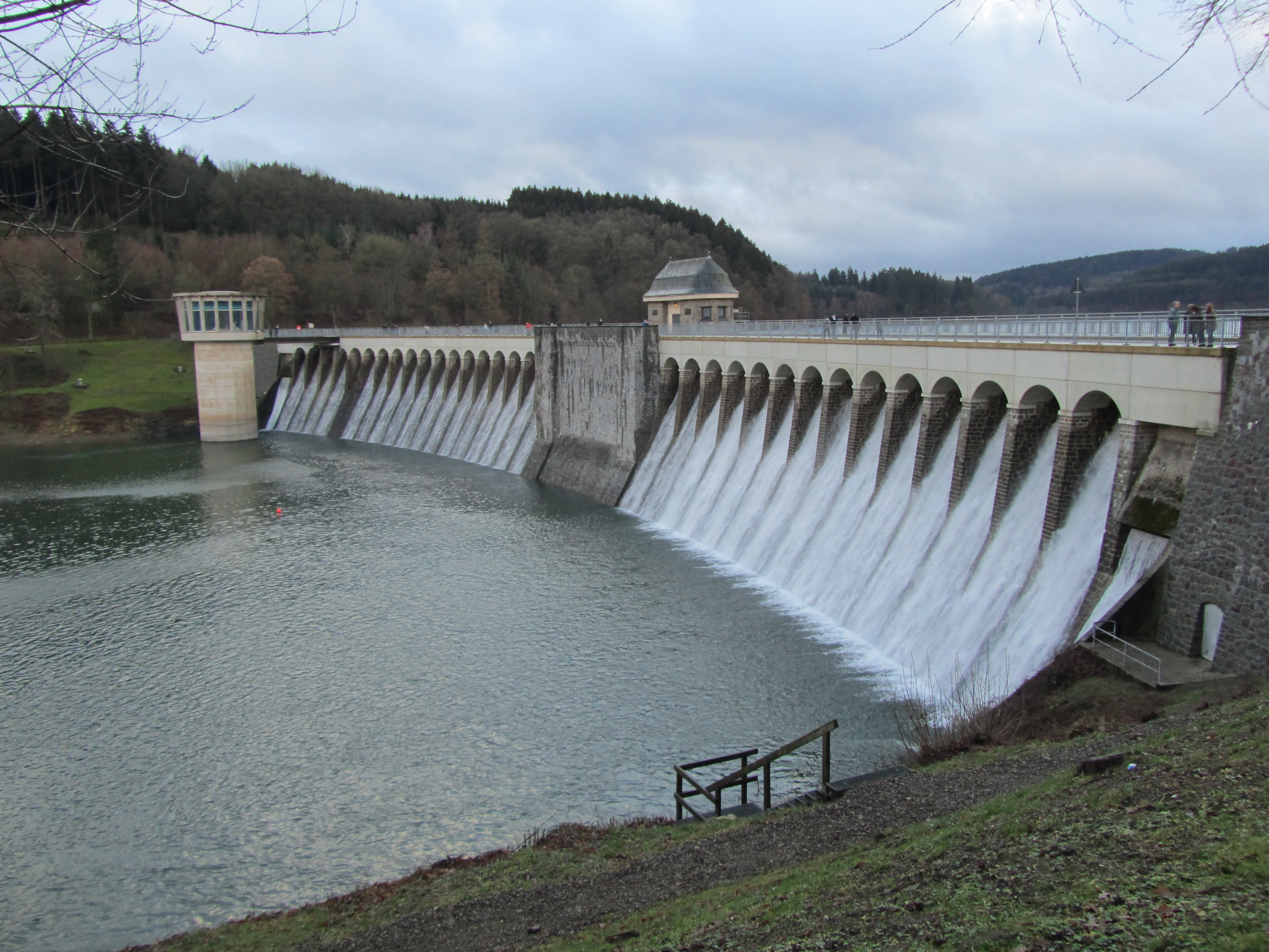 A stone dam spillway with multiple arched openings, releasing cascading water into the reservoir behind a forested hillside under an overcast sky.