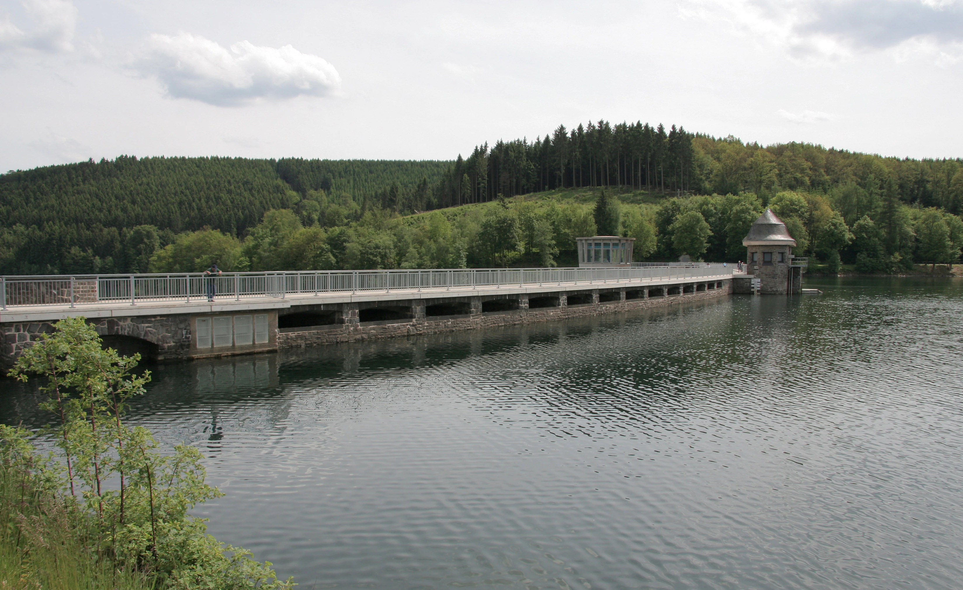 A wide angle view of the concrete dam wall stretching across the Listertalsperre reservoir, with a small stone tower on the right and forested hills in the background.