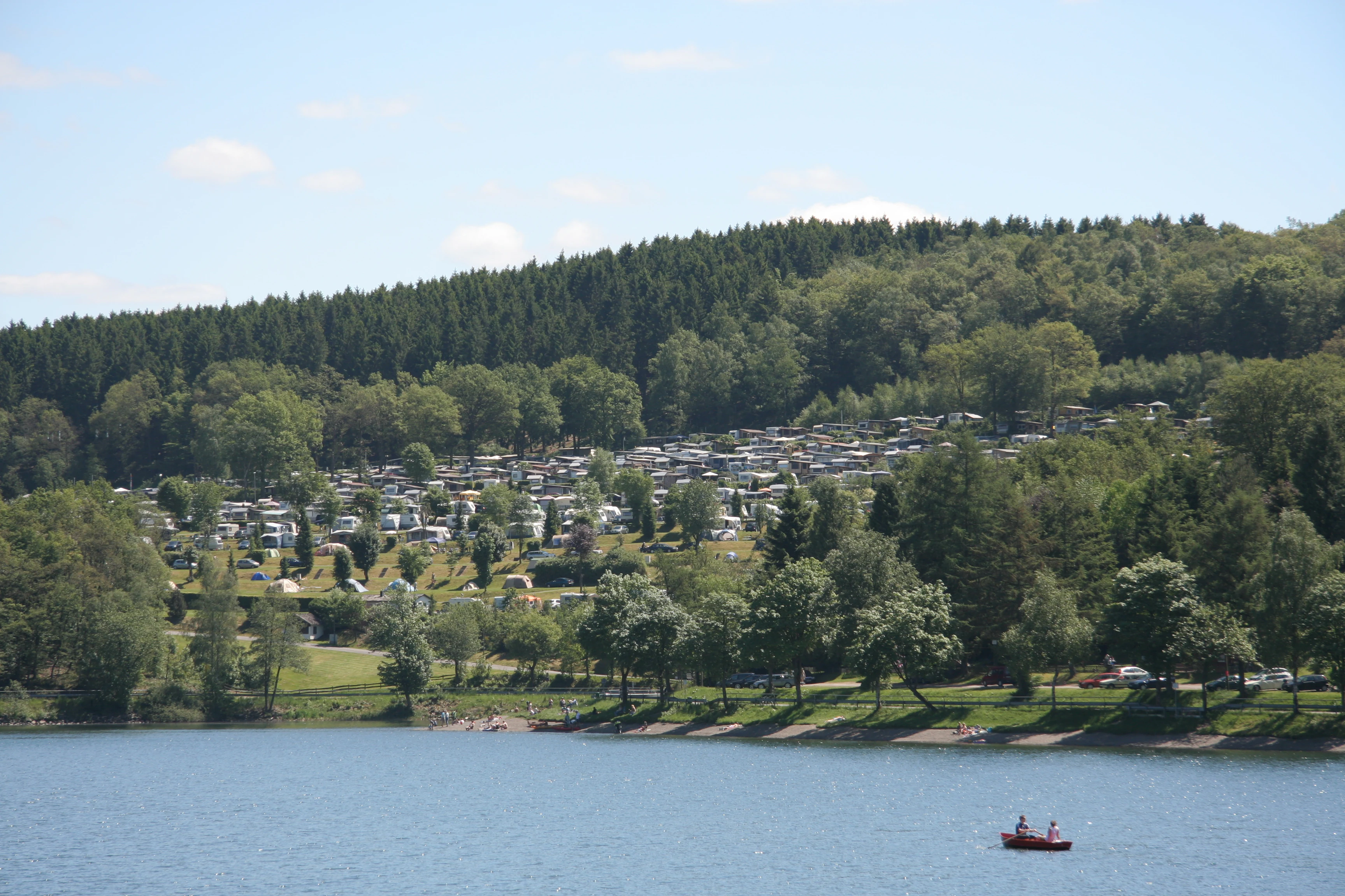 A wide shot showing a large campground nestled into a forested hillside overlooking the Listertalsperre reservoir, with a small boat visible on the water.