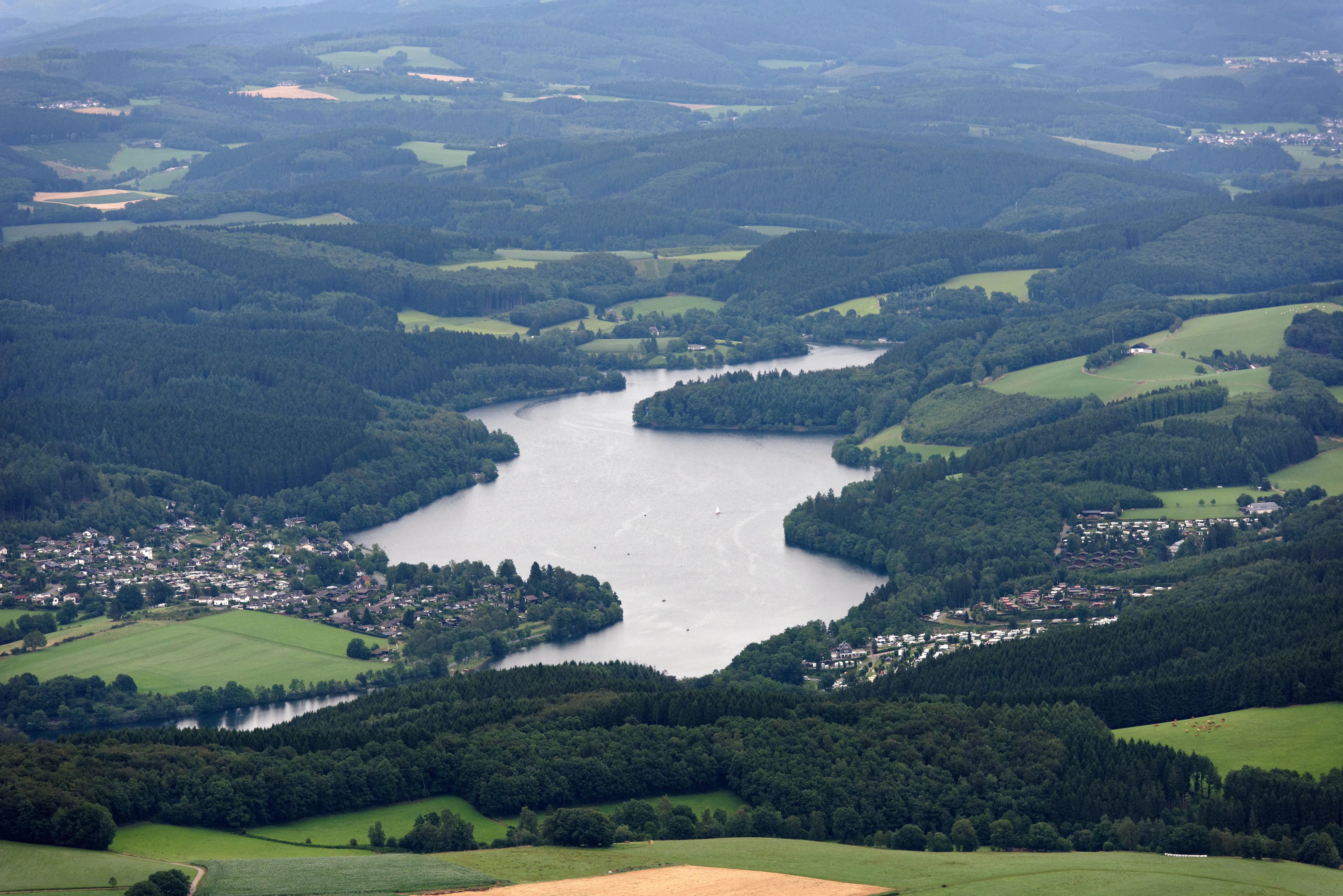An aerial view of the Listertalsperre reservoir, surrounded by lush green forests, grassy hills, and residential areas.