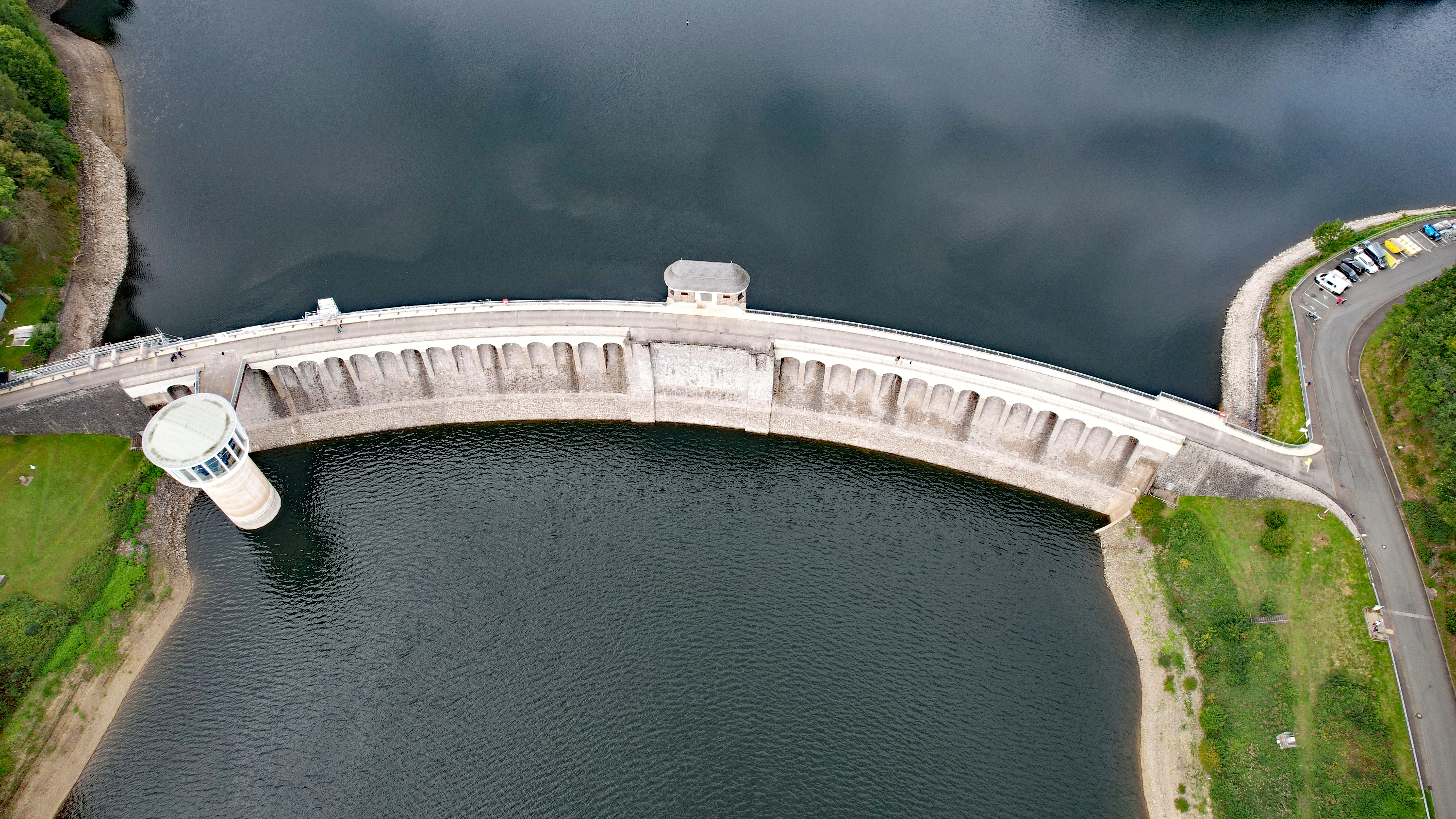 An overhead aerial view of a curved concrete dam wall separating two bodies of dark water, with a small observation tower in the water and a road leading to the bank.