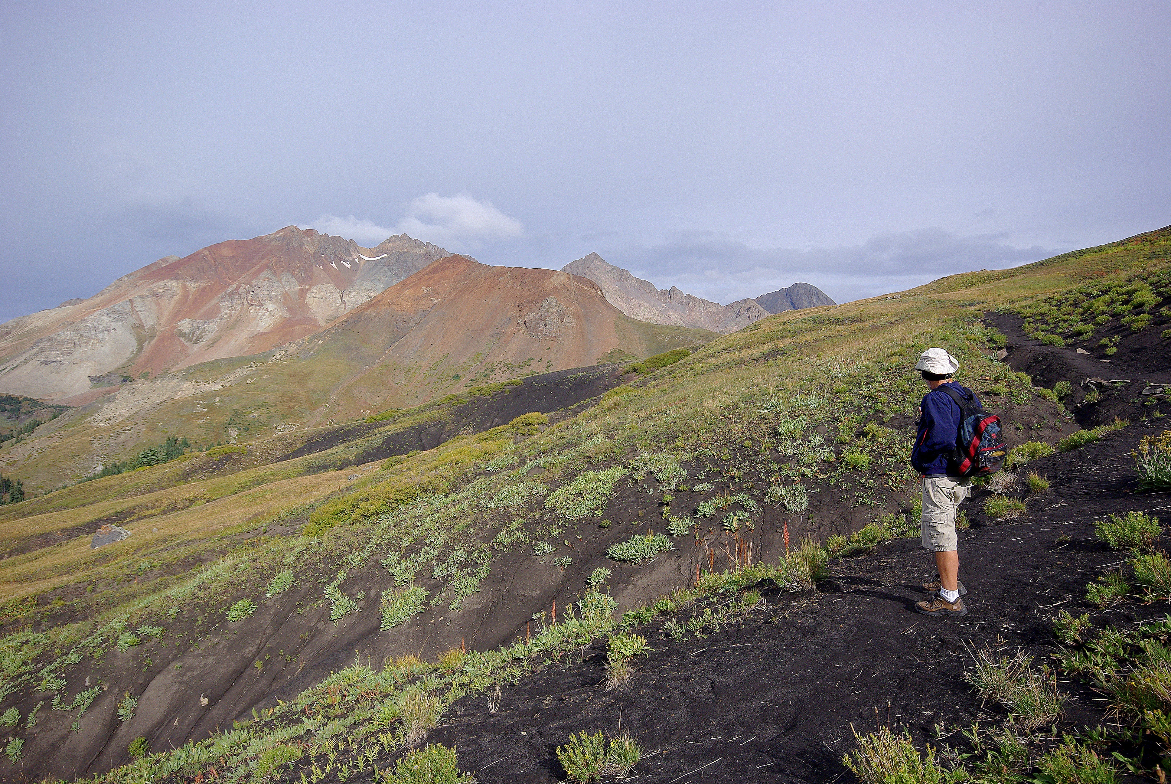 A hiker with a backpack stands on a dark, rocky trail overlooking a sweeping mountain landscape with colorful volcanic slopes.