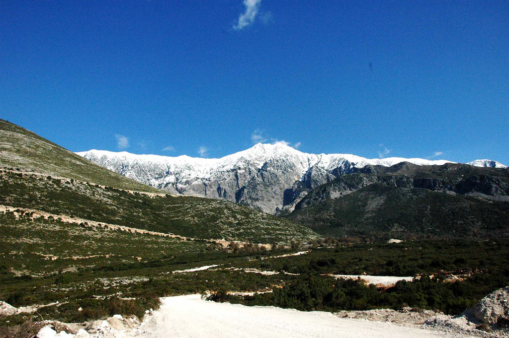 Snow-capped mountain peaks under a clear blue sky with a dirt road and rolling hills in the foreground