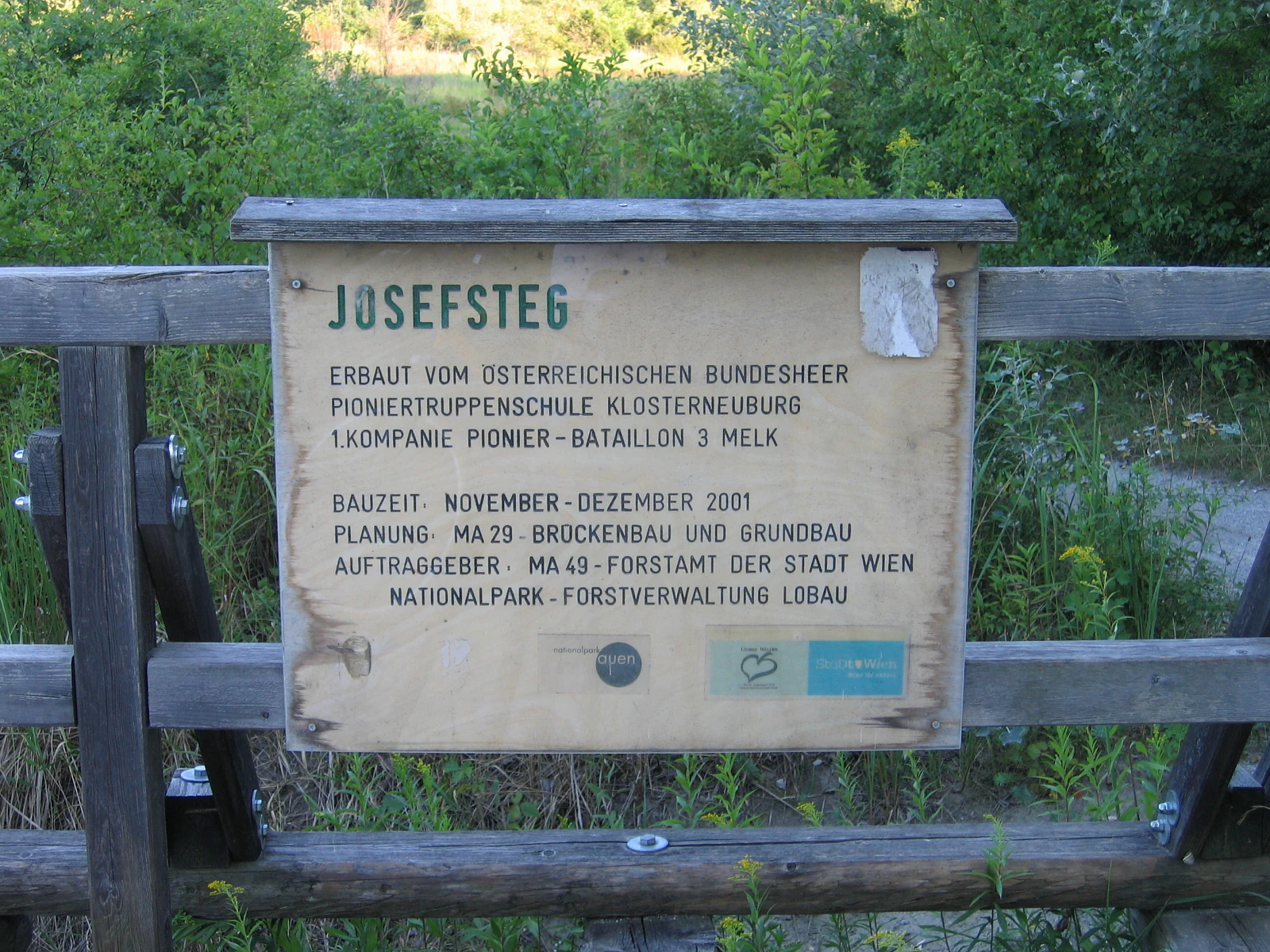 A rectangular wooden information sign mounted on a rustic wooden railing, displaying text about the Josefsteg bridge and its construction history against a blurred background of green foliage.