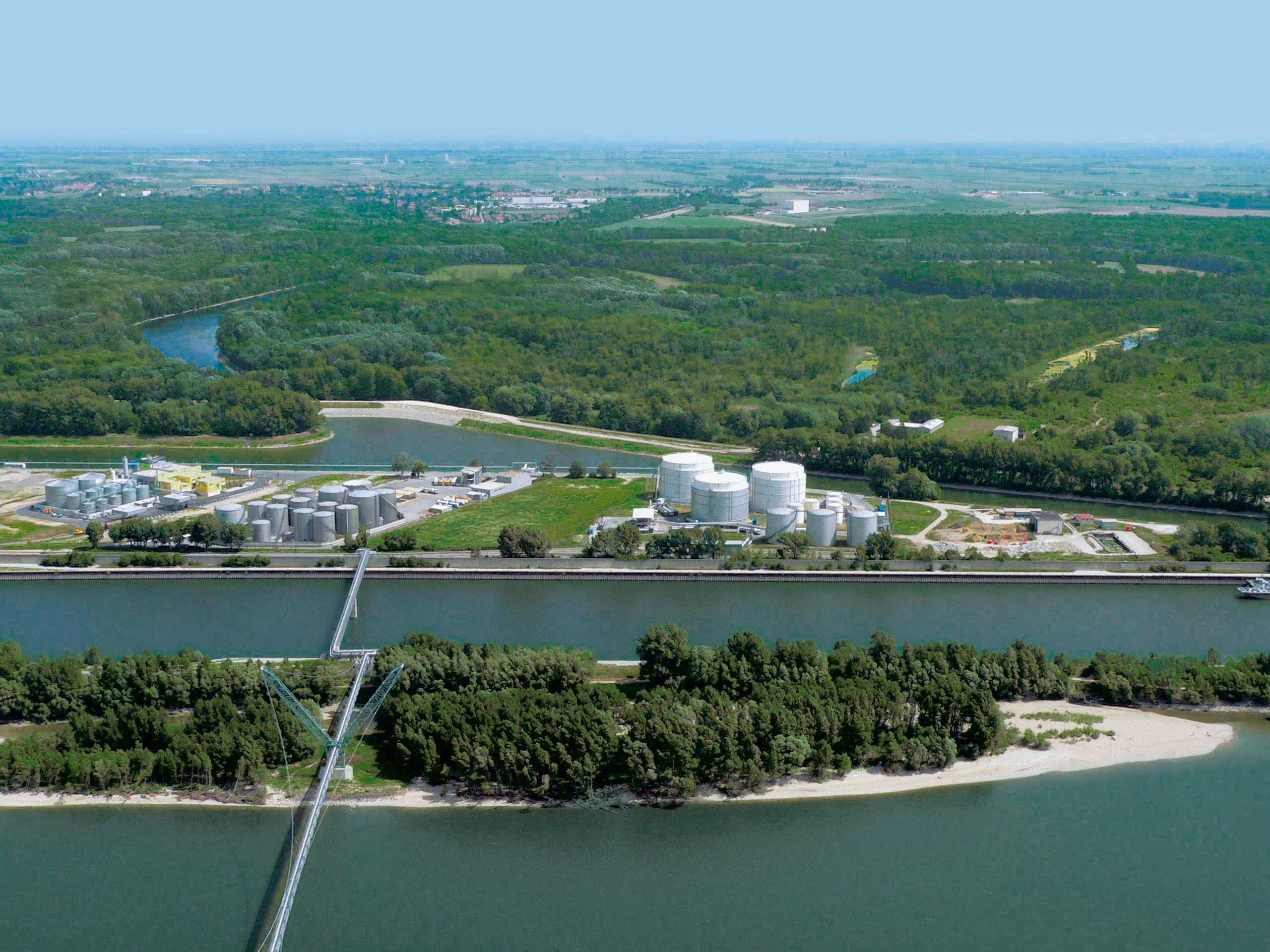 Aerial view looking over a wide river, a narrow canal with a pedestrian bridge, and a forested area containing large industrial storage tanks.