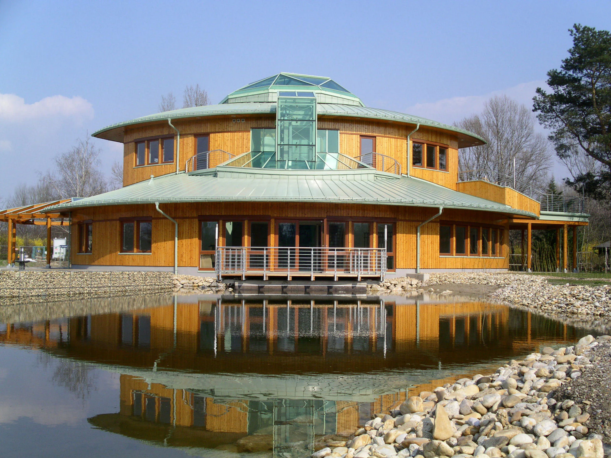 A modern, two-story, circular wooden visitor center building with a green metal roof, reflecting in a calm pond in the foreground.