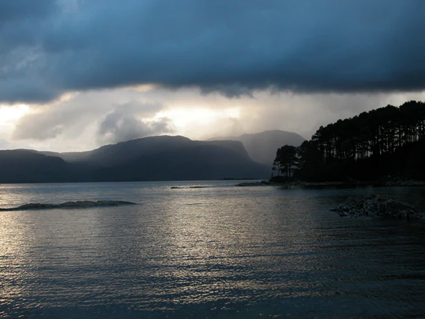 A wide, scenic view of Loch Carron with dark, cloudy skies above, silhouettes of hills in the distance, and a treelined shoreline in the midground.
