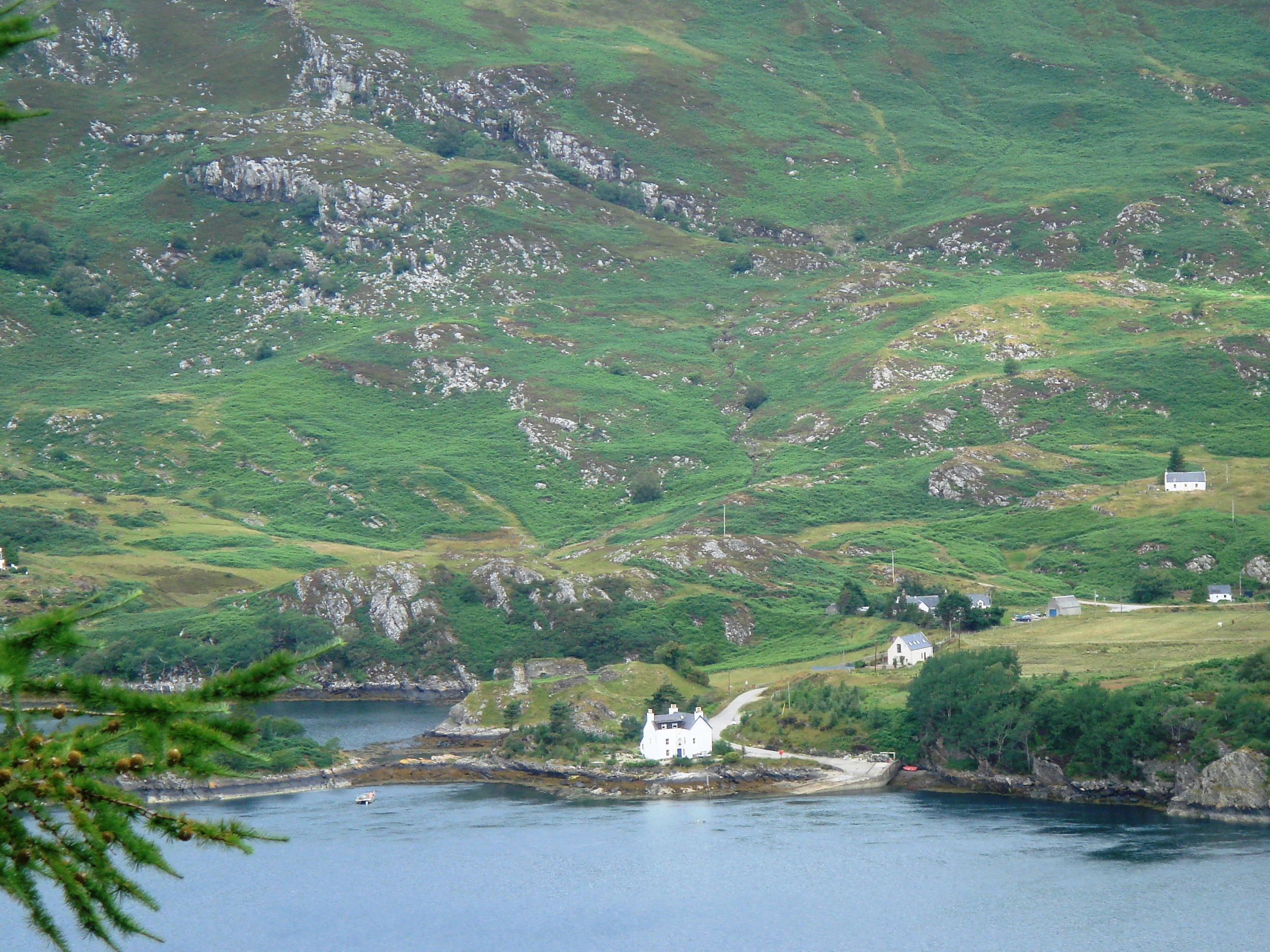 A high-angle view of Loch Carron, featuring calm dark water, a small white building situated on a low promontory near the shore, and rolling green, hilly terrain with exposed rocky patches in the background.