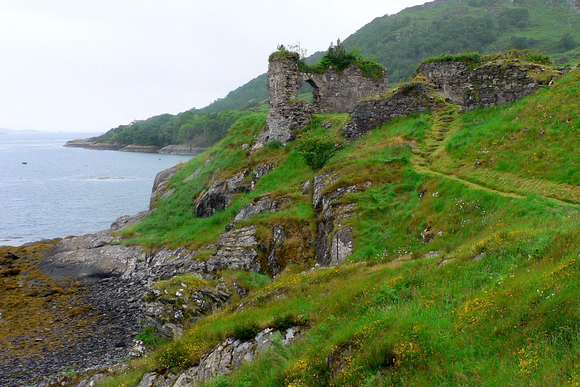 A stone castle ruin stands on a grassy, rocky slope overlooking a calm body of water under an overcast sky.