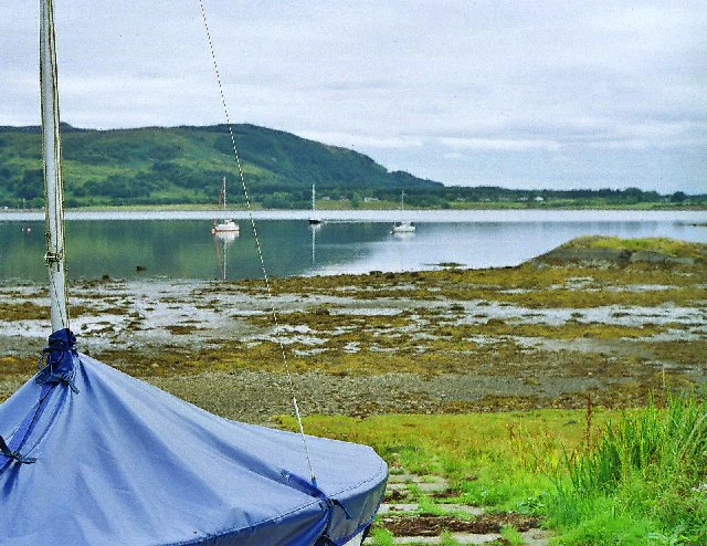 A covered sailboat sits in the foreground on a grassy bank at low tide, with the calm water of a sea loch and distant green hills under an overcast sky in the background.