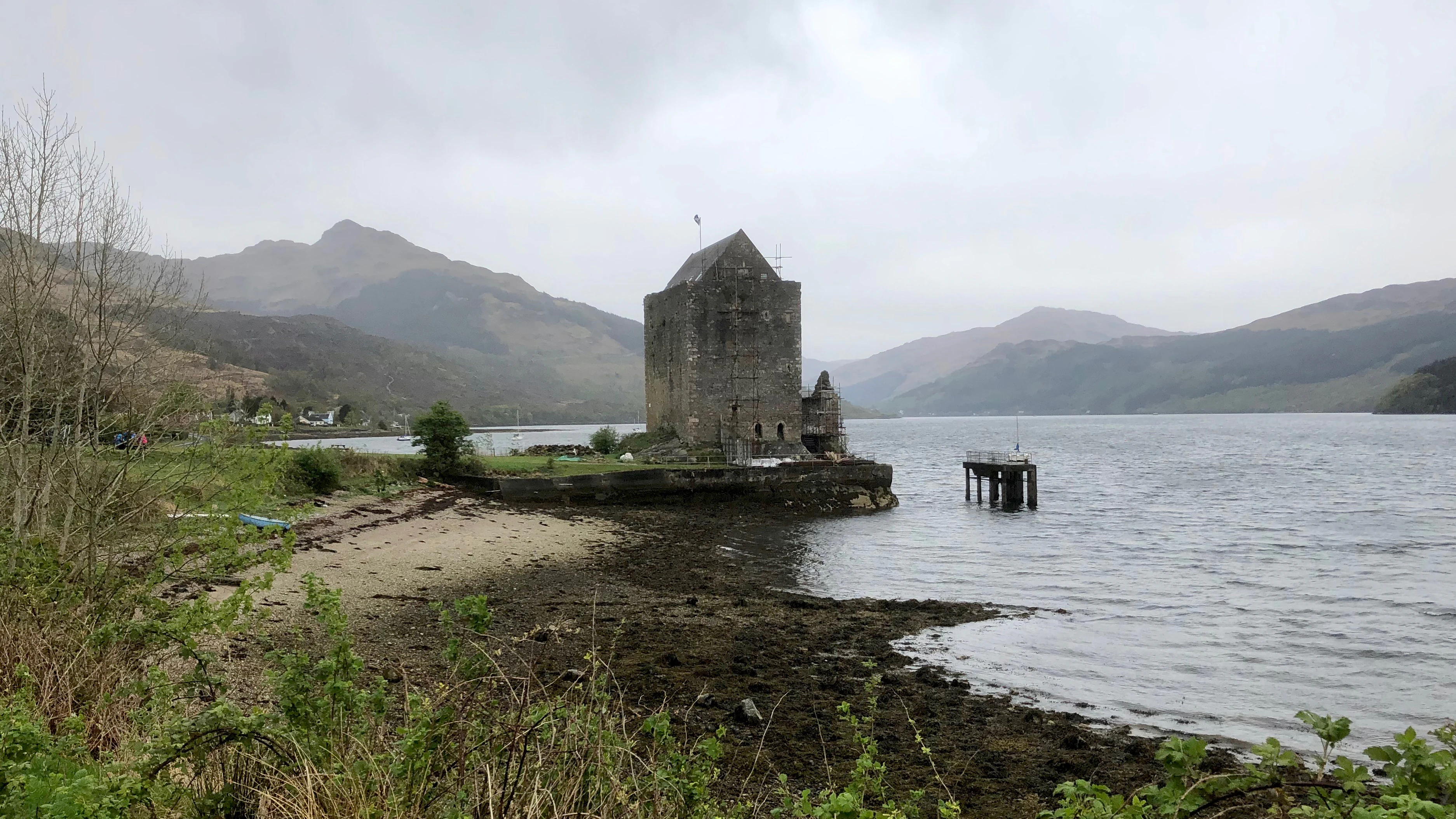 A stone tower house, Carrick Castle, sits on a small promontory extending into the dark waters of Loch Goil, surrounded by rolling hills under a cloudy sky.