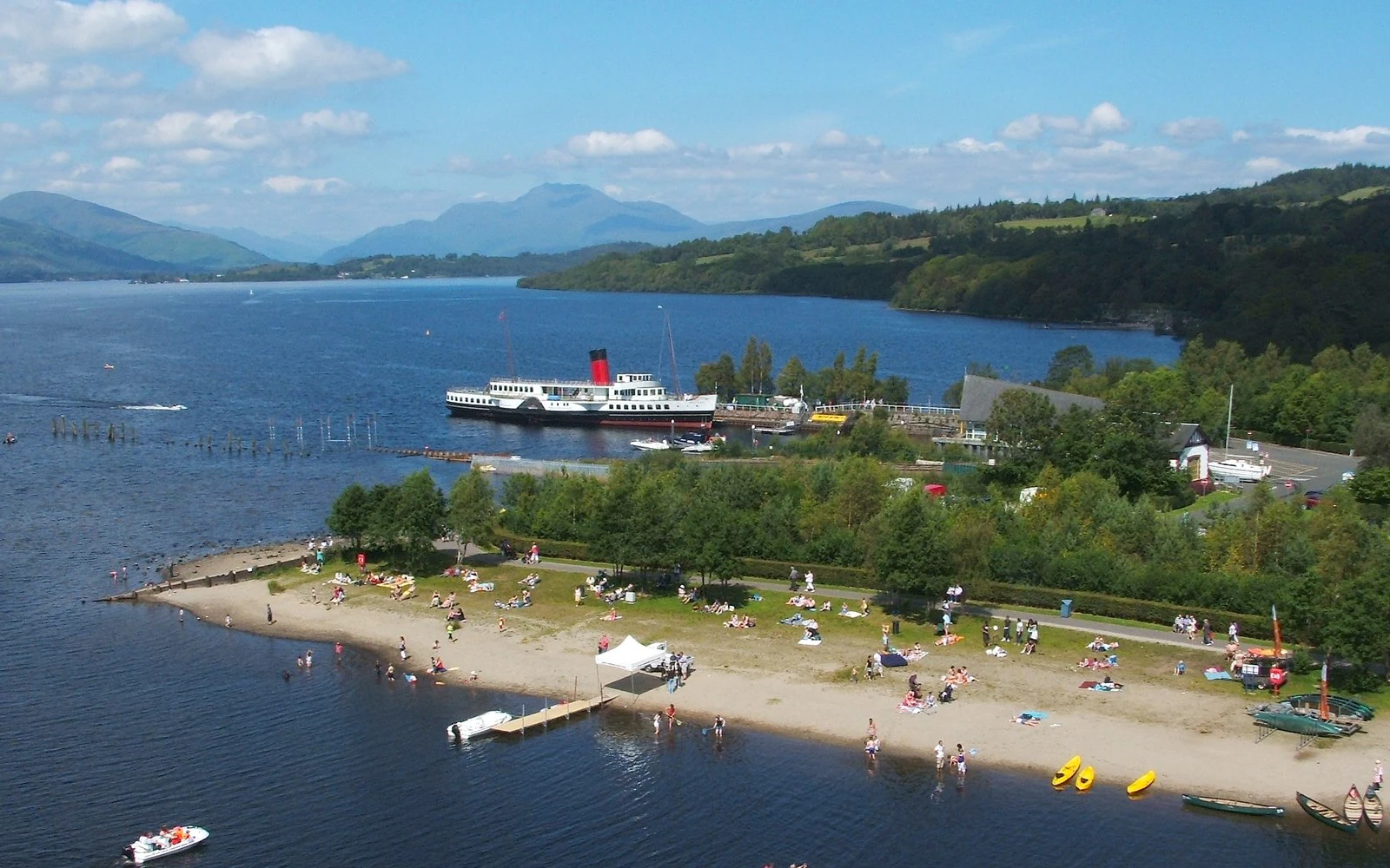 An elevated view of a sandy lakeshore with people, small boats, and a large historic ferry docked near a treed shoreline with mountains in the background.