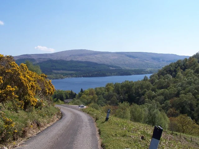 A winding asphalt road in the foreground leads toward a large blue lake surrounded by rolling green hills and mountains under a clear blue sky.