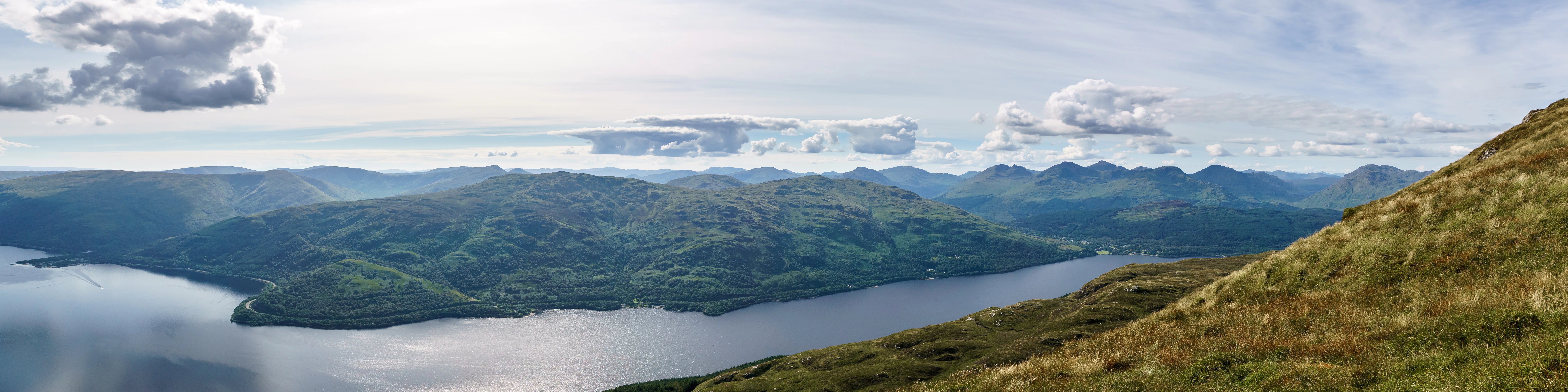 A wide, panoramic, eye-level landscape shot featuring a large, dark blue lake winding through rolling green hills and distant mountain ranges under a partly cloudy sky.