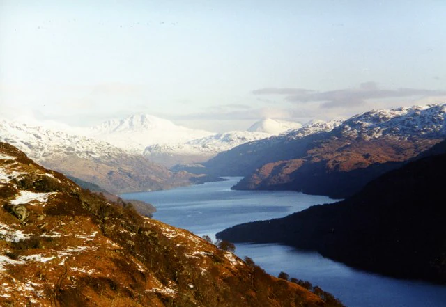 A wide angle view of a large, long lake stretching through a valley, flanked by snow-dusted mountains and rugged highland hills.