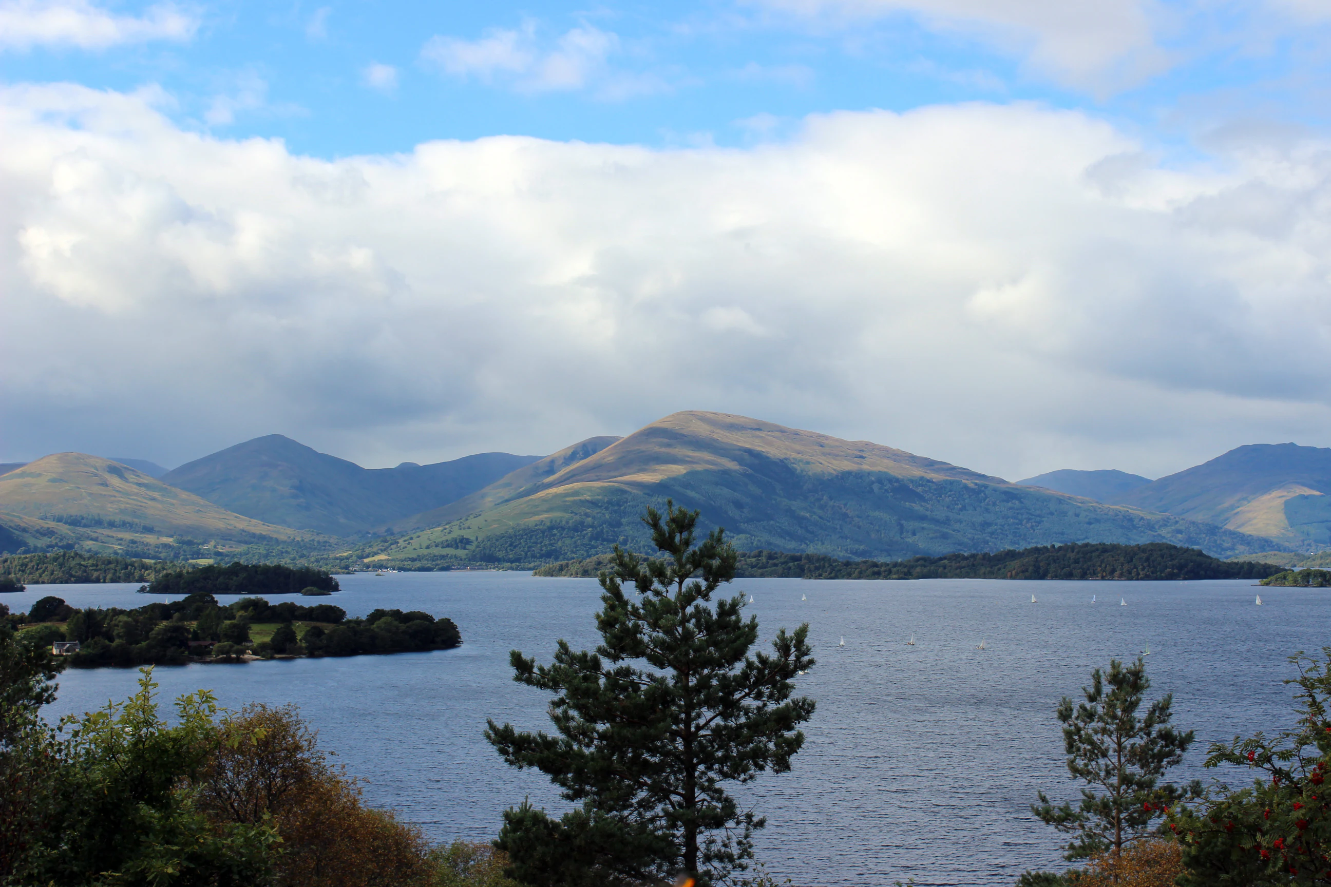 A wide shot of Loch Lomond with small forested islands in the water, framed by distant rolling mountains under a cloudy sky.