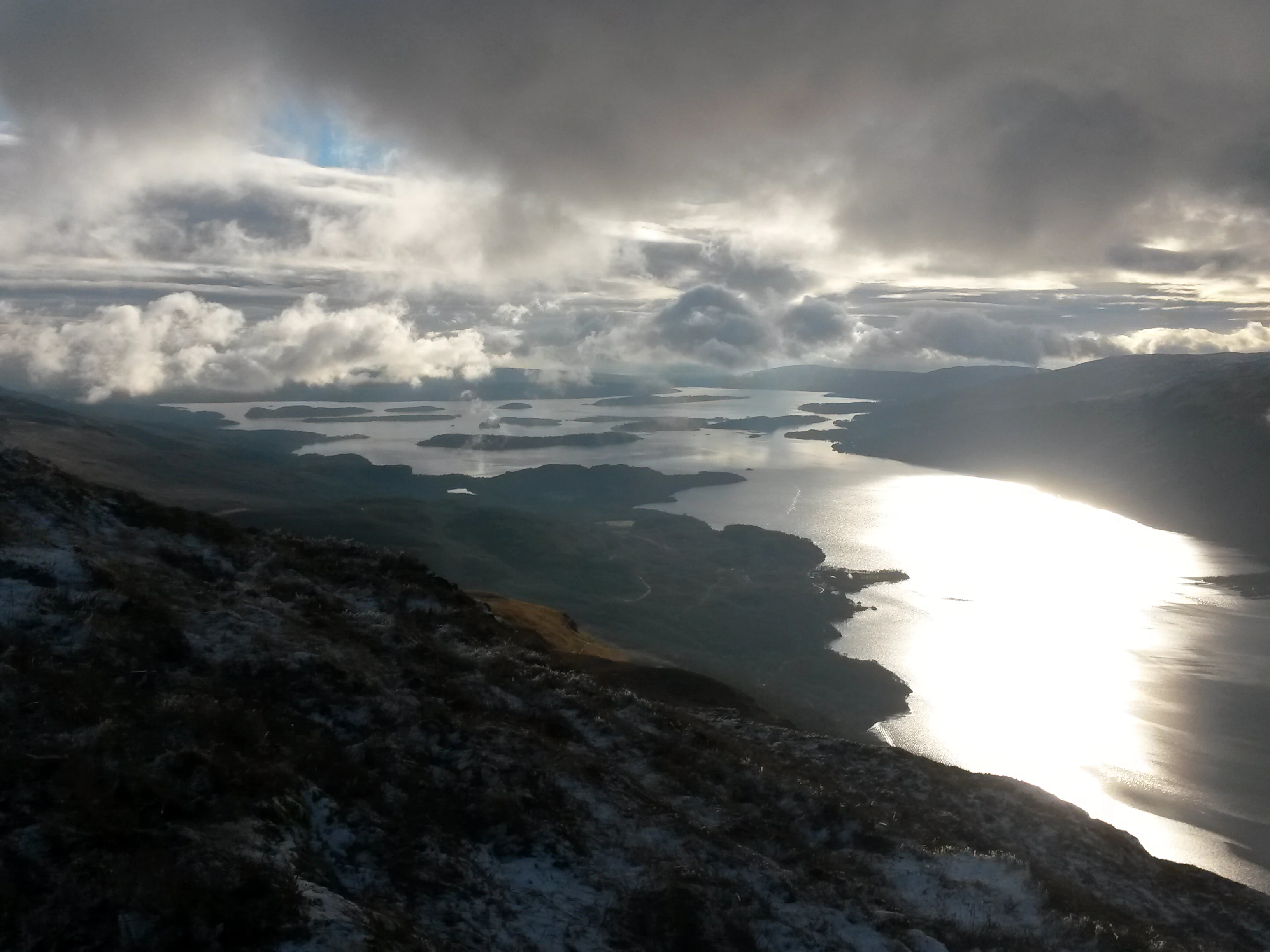 Panoramic view of Loch Lomond with mountains in the background under a partly cloudy sky, sunlight reflecting on the water