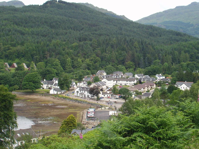 A village with houses and trees surrounded by hills and a body of water, viewed from a high vantage point