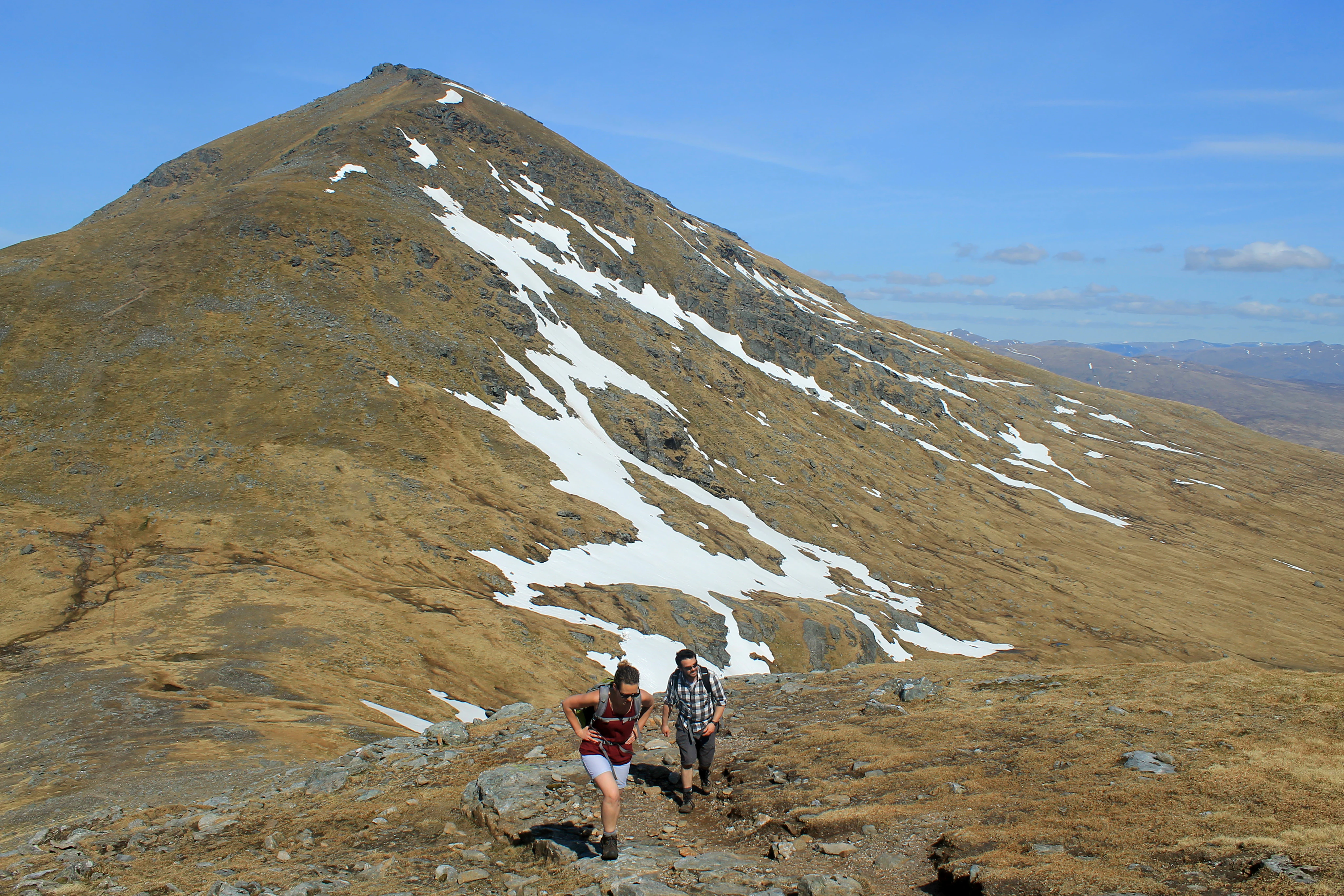 Two hikers on a rocky mountain ridge with Ben More mountain featuring snow patches in the background under a clear blue sky