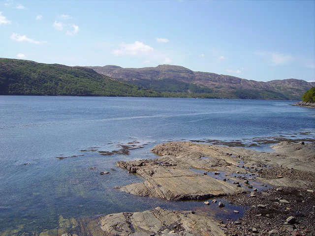 A wide view of a calm sea loch with rocky shoreline in the foreground and rolling hills in the background under a blue sky.
