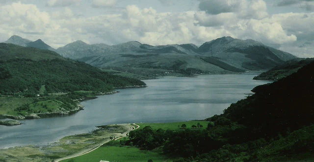 A wide, elevated landscape view of a calm sea loch surrounded by rolling green hills and rugged mountains under a cloudy sky.